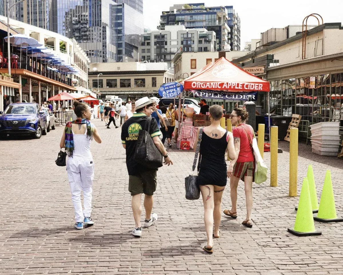 شخصيات متنوعة تتجول في سوق بايك بلايس في سياتل، مع أكشاك الطعام والمباني الحديثة في الخلفية، تعكس انخفاض عدد الزوار الكنديين.