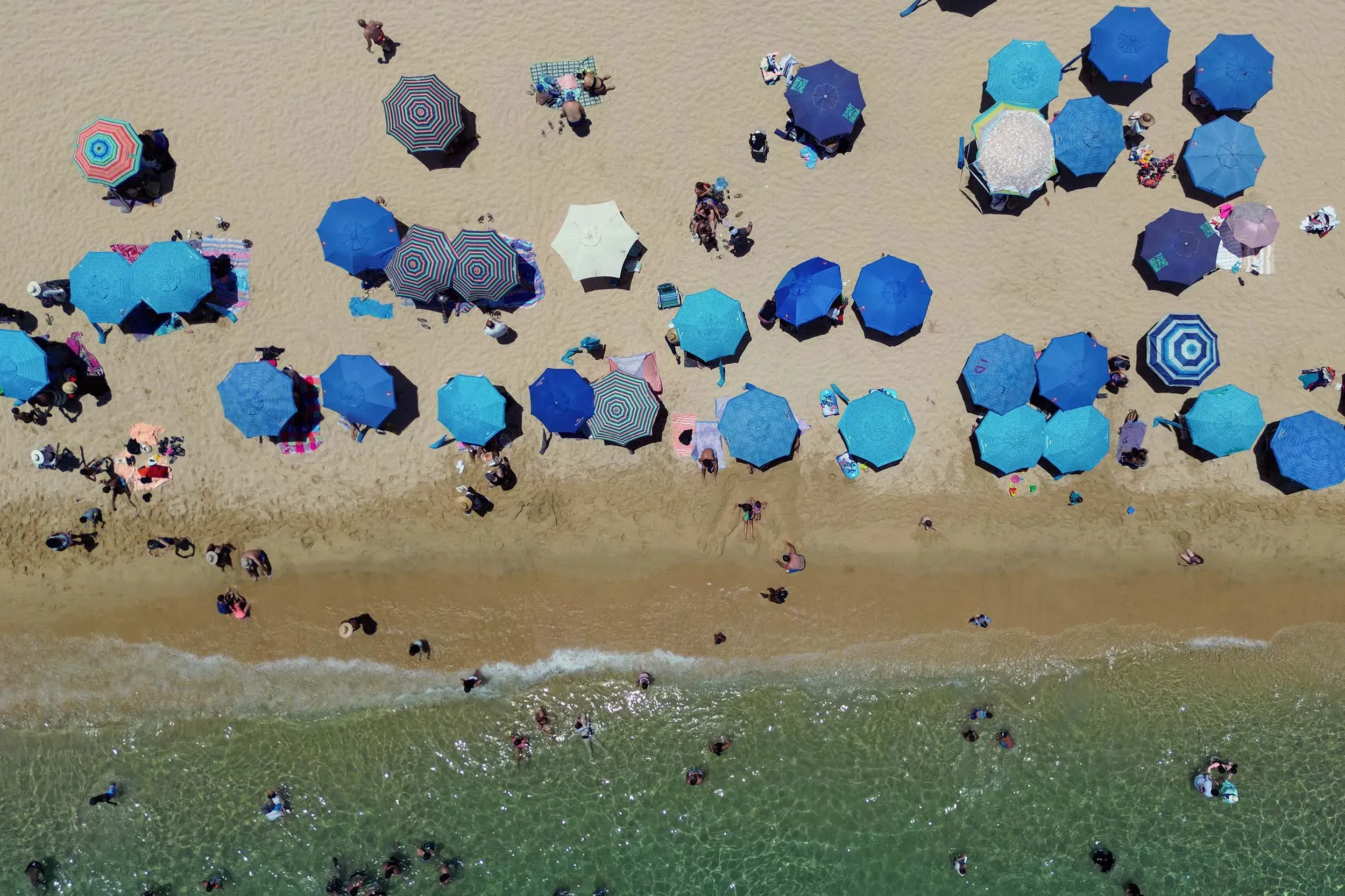 شاطئ مزدحم بالأشخاص تحت مظلات زرقاء متعددة الأشكال، مع أمواج البحر تتلاطم على الشاطئ، مما يعكس تأثير انخفاض السياحة.