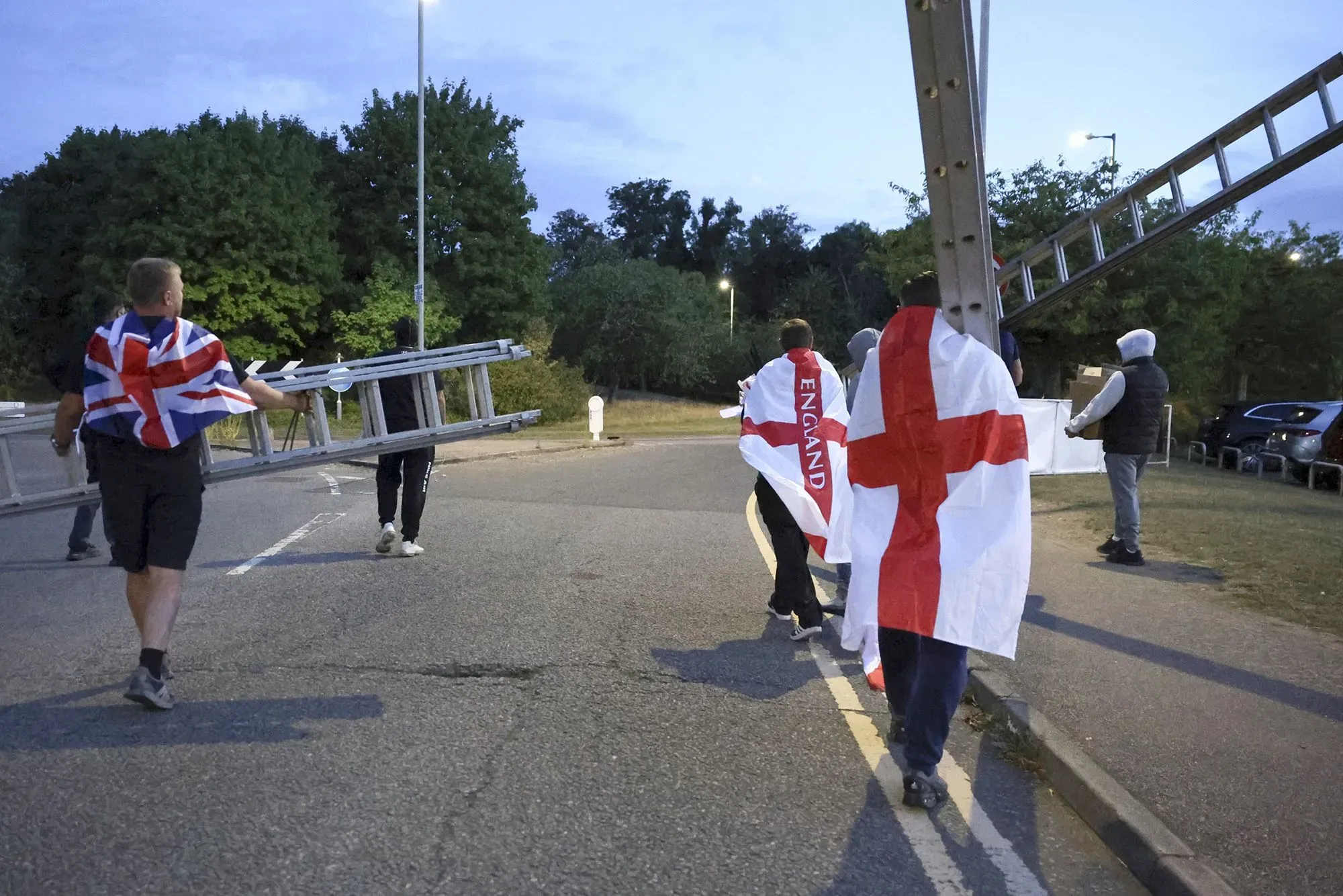 مجموعة من الرجال يحملون سلالم ويرتدون أعلام إنجلترا والاتحاد، يتجهون نحو موقع لرفع الأعلام في الشارع.