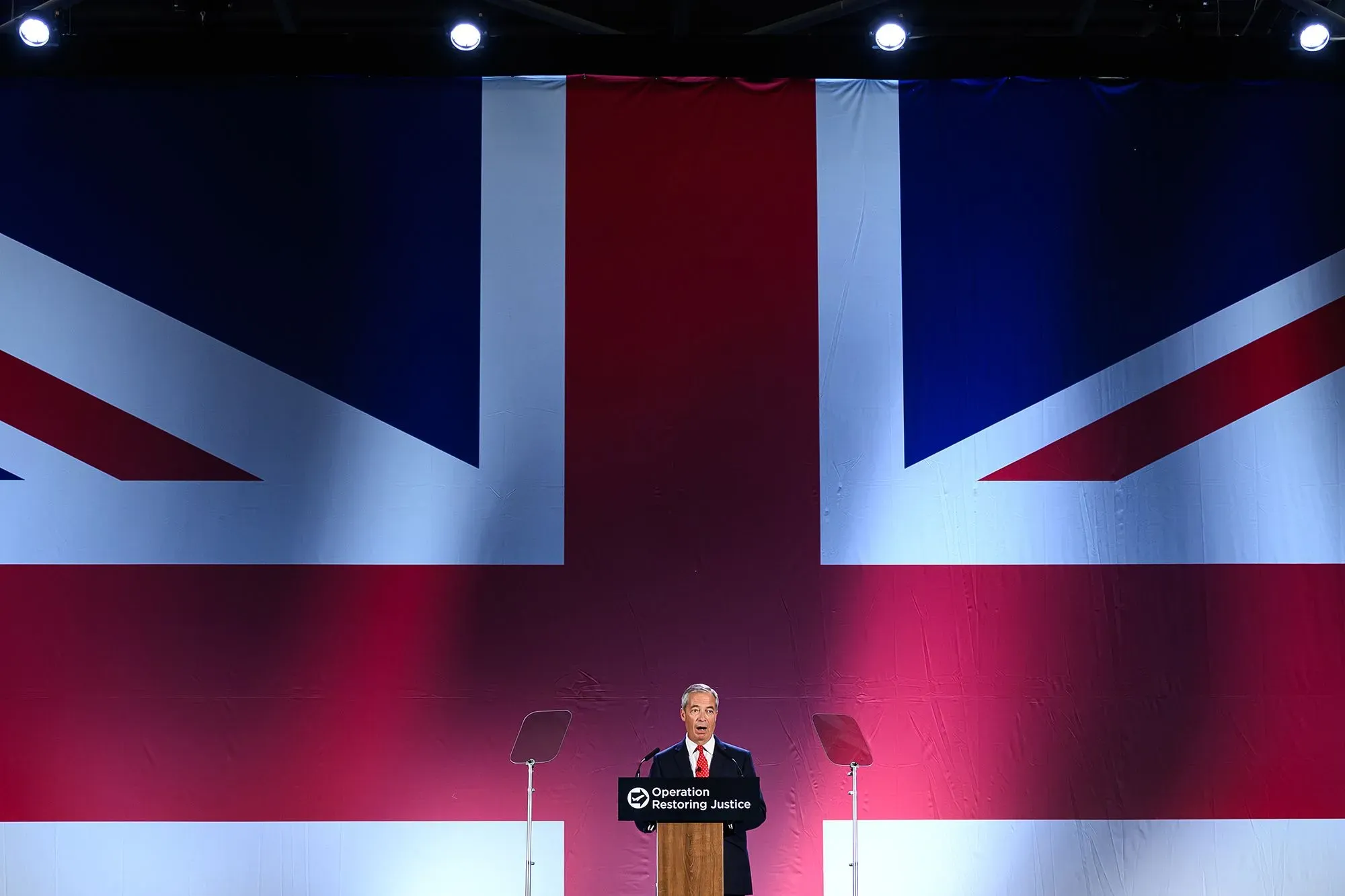 تظهر الصورة منصة يتحدث عليها نايجل فاراج، مع خلفية كبيرة لعلم المملكة المتحدة. تعكس الأعلام الوطنية المشاعر الوطنية المتزايدة في إنجلترا.