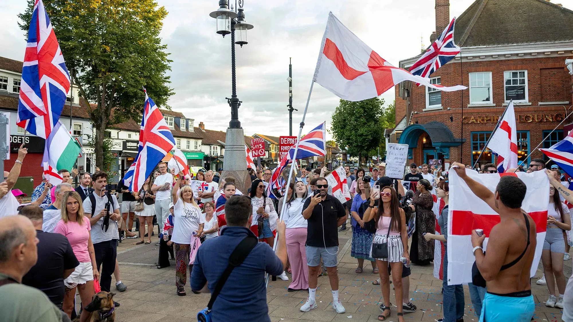 تجمع حشد من الناس في الشارع يحملون أعلام المملكة المتحدة وعلم إنجلترا، مع شعارات تعبر عن مشاعر وطنية في سياق سياسي مشحون.