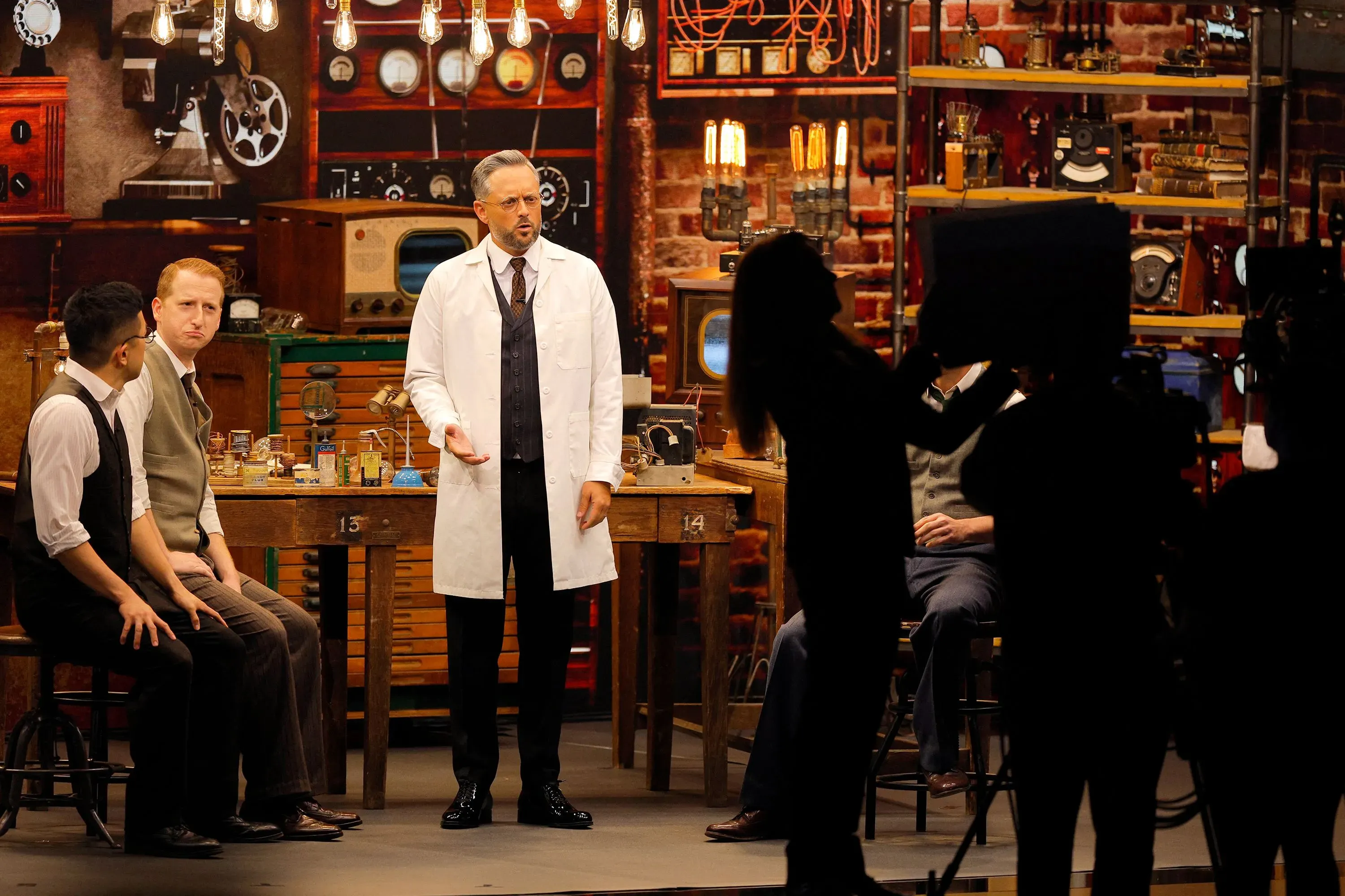 مشهد من عرض "ذا ستوديو" حيث يظهر الممثلون في ملابس تاريخية أثناء تصوير مشهد، مع معدات تصوير وخلفية تحتوي على أدوات قديمة.