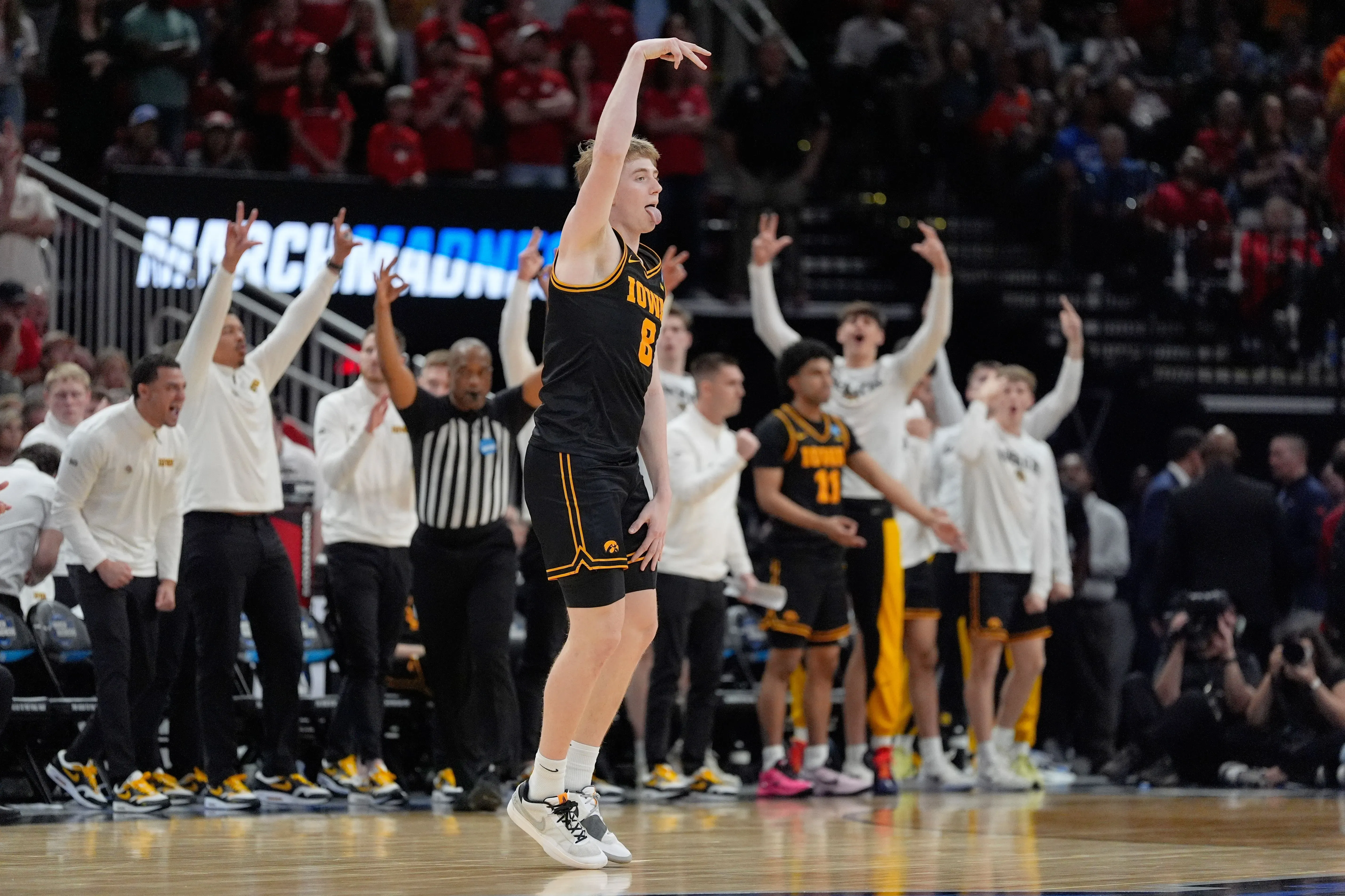 لاعب كرة سلة من ولاية أيوا يحتفل بتسجيل نقطة، بينما يشجع زملاؤه في الفريق على خلفية حماسية خلال مباراة بطولة جنون مارس.