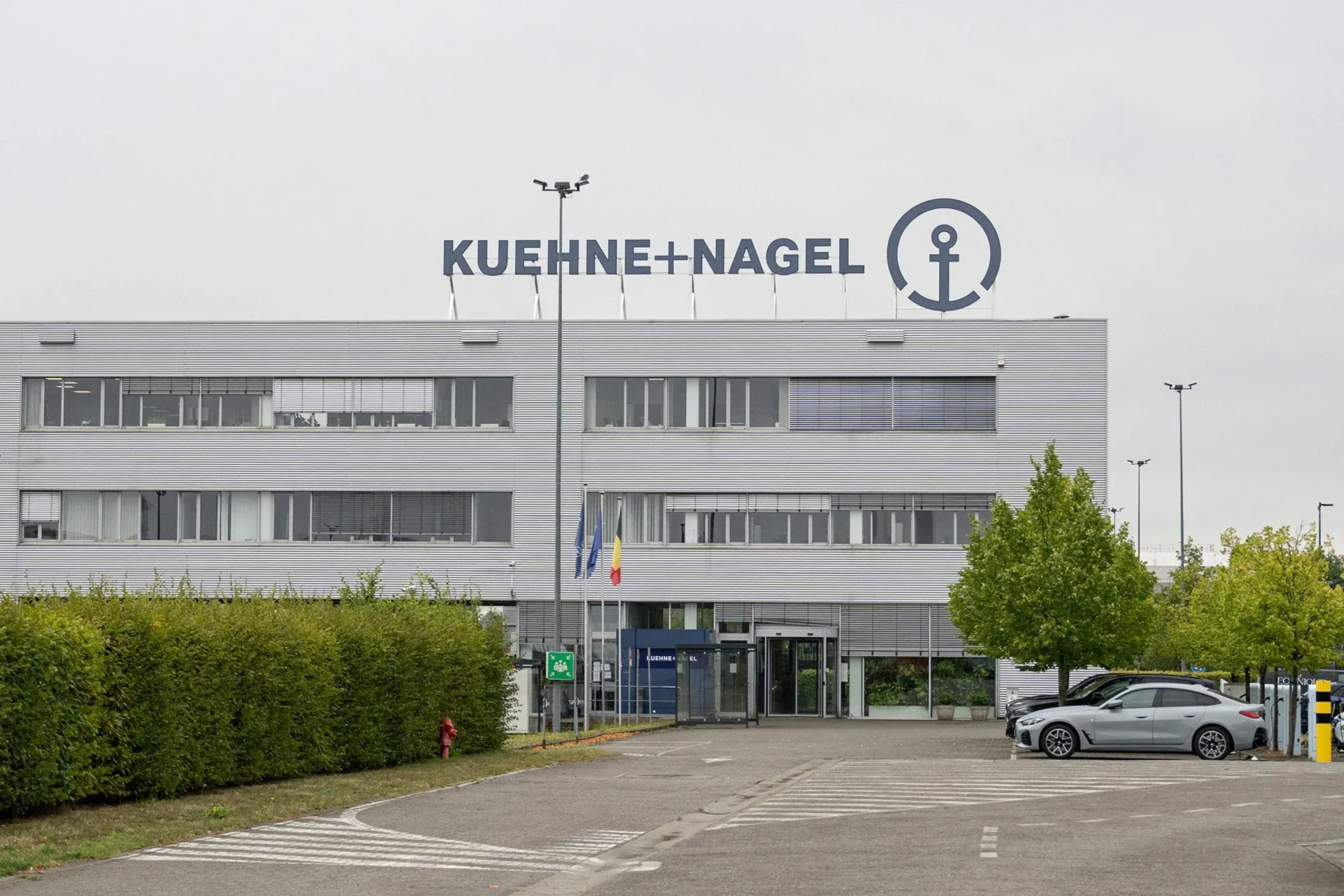 مبنى شركة Kuehne + Nagel يظهر في الصورة، مع لافتة الشركة واضحة على السطح، ويحيط به مساحات خضراء ومواقف للسيارات.