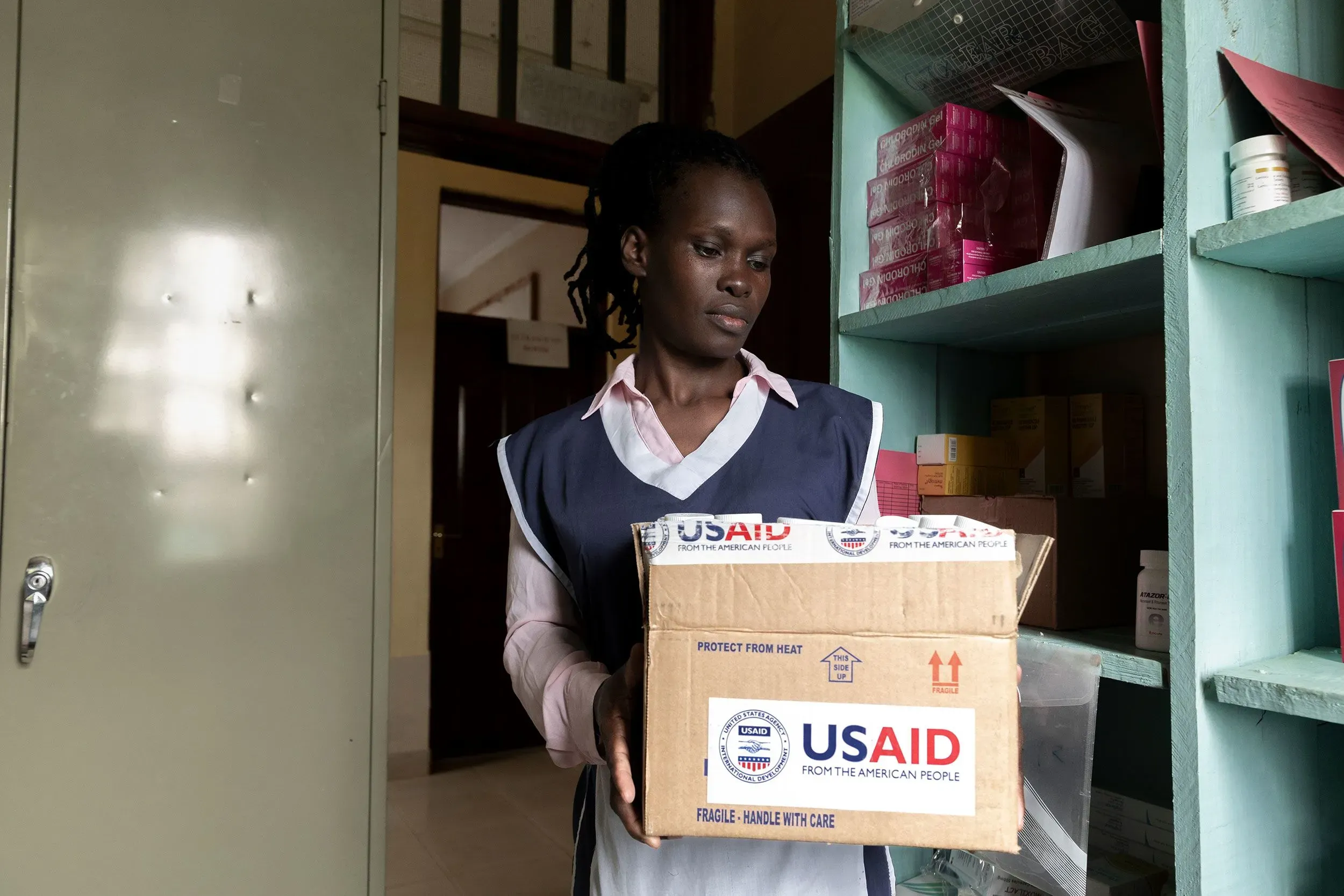 امرأة تحمل صندوقًا يحمل شعار الوكالة الأمريكية للتنمية الدولية (USAID)، في مركز صحي، مما يعكس تأثير التمويل على خدمات الإغاثة من فيروس نقص المناعة البشرية.