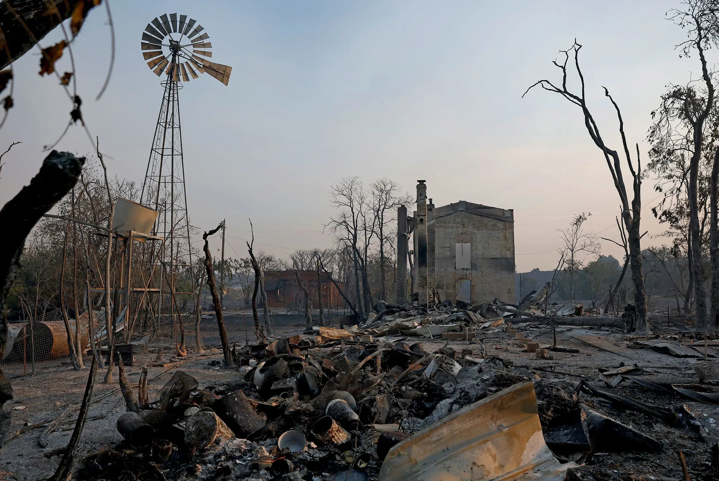 منظر لخراب ناجم عن حرائق الغابات، حيث تظهر أنقاض مبنى متضرر، وأشجار محترقة، وعجلات مائية في الخلفية، مما يعكس آثار الكوارث الطبيعية.