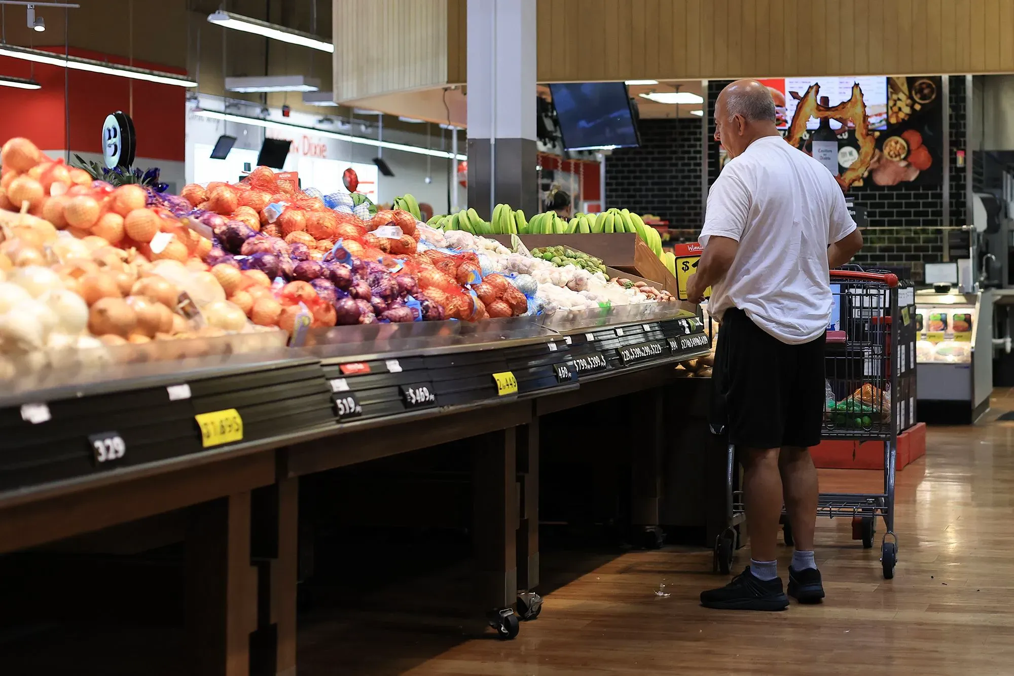 رجل يتسوق في سوبرماركت، محاط بخضروات وفواكه متنوعة، مما يعكس تأثير ارتفاع أسعار الغذاء بسبب إغلاق مضيق هرمز.