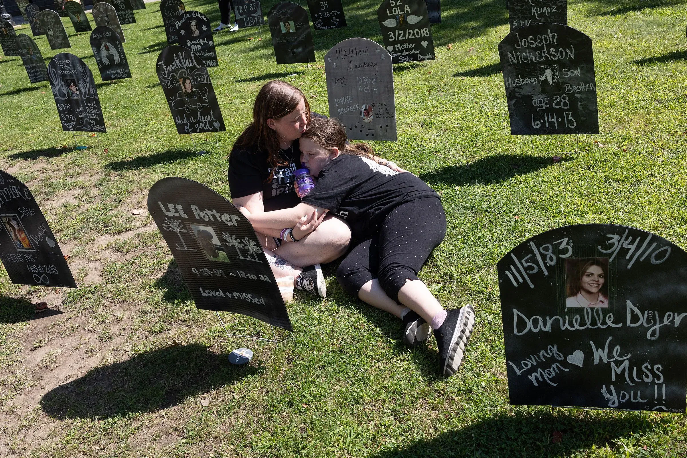 امرأتان تجلسان على العشب بين شواهد قبور صغيرة تحمل أسماء ضحايا المخدرات، تعبير عن الحزن والفقد.