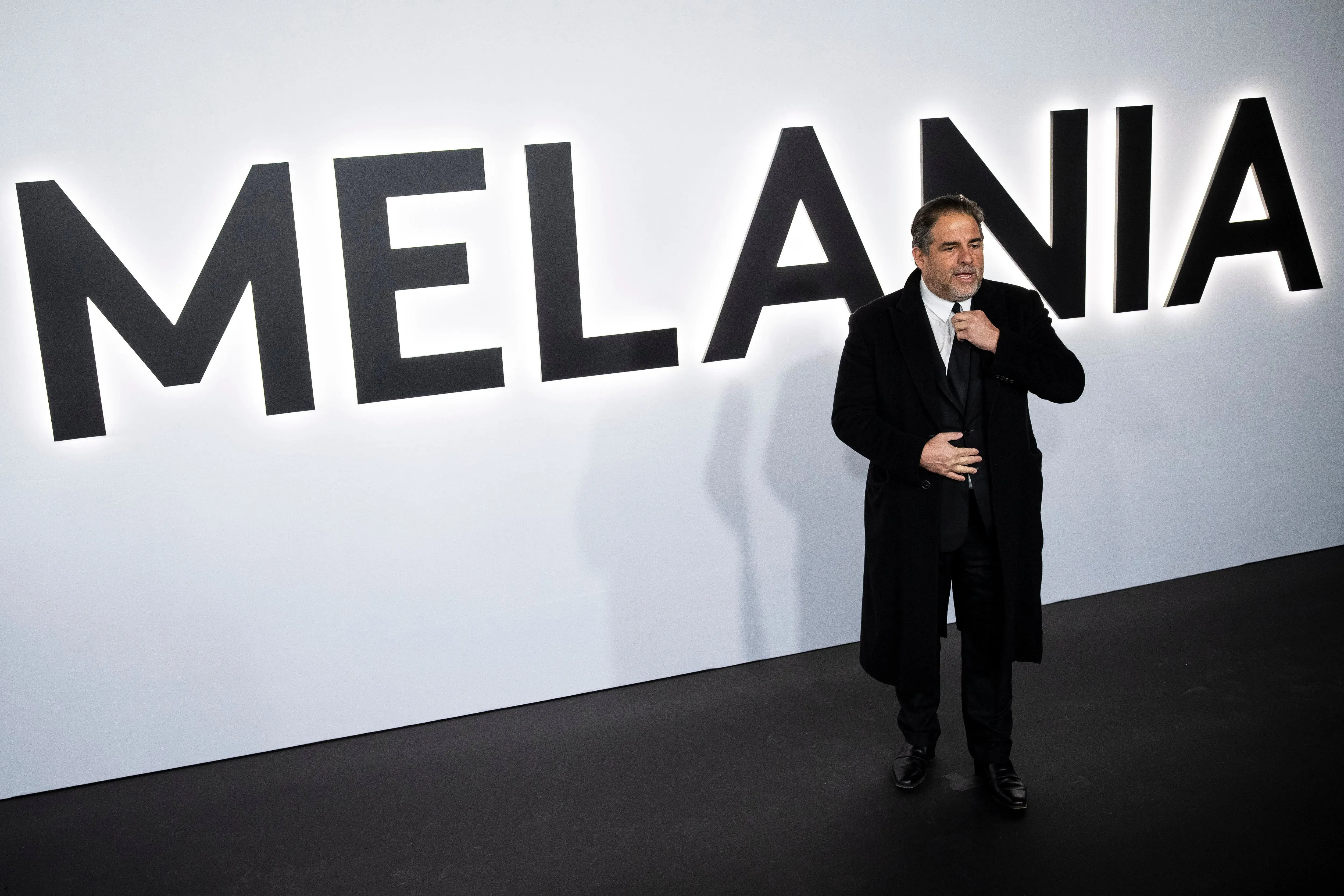 رجل يرتدي بدلة سوداء يتحدث أمام خلفية تحمل اسم "ميلانيا" بخط عريض، مما يشير إلى حدث أو عرض متعلق بها.