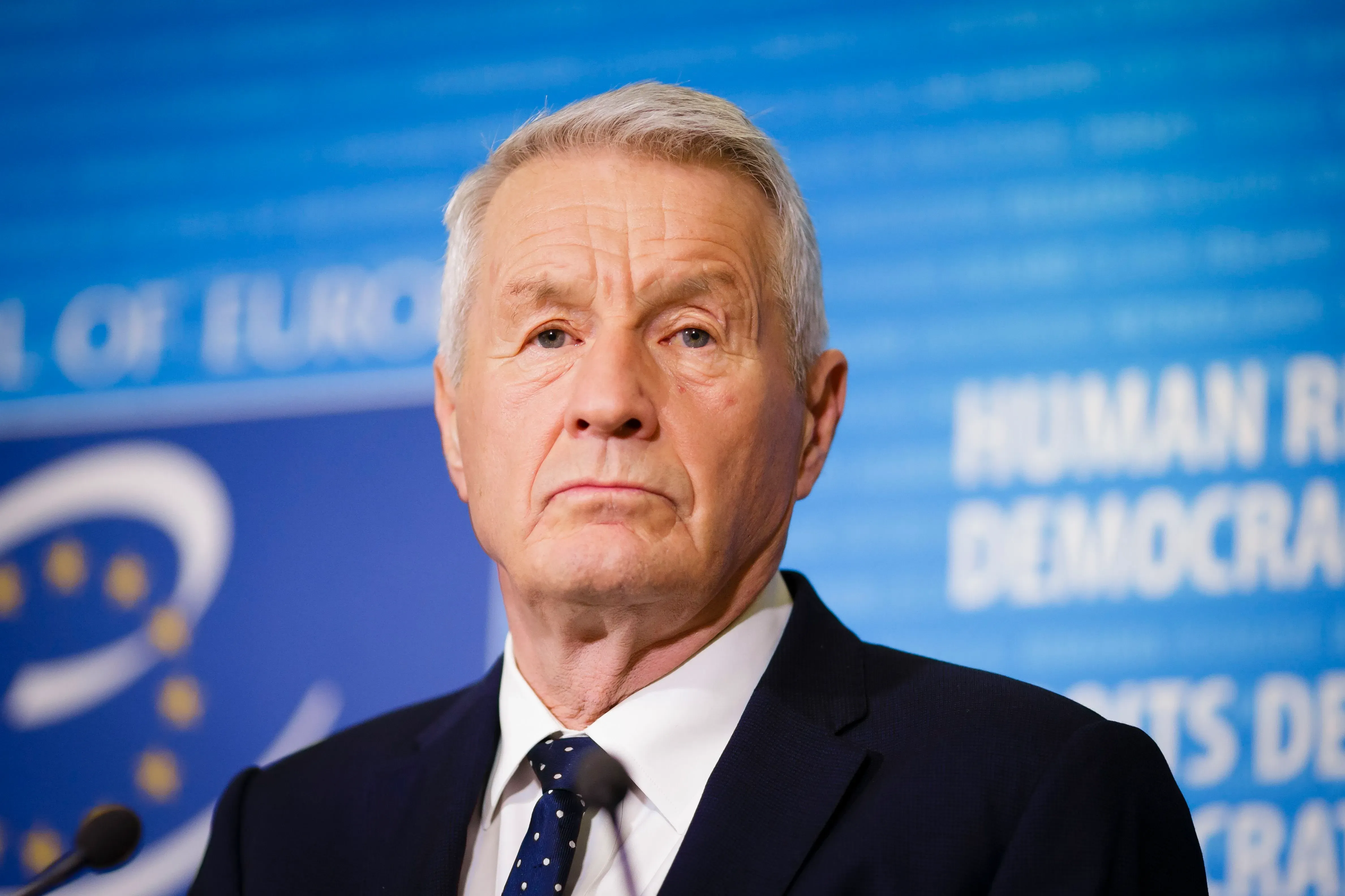 صورة لرجل مسن يرتدي بدلة رسمية، يبدو جادًا ويظهر تعبيرًا حازمًا، مع خلفية تحمل شعار حقوق الإنسان.