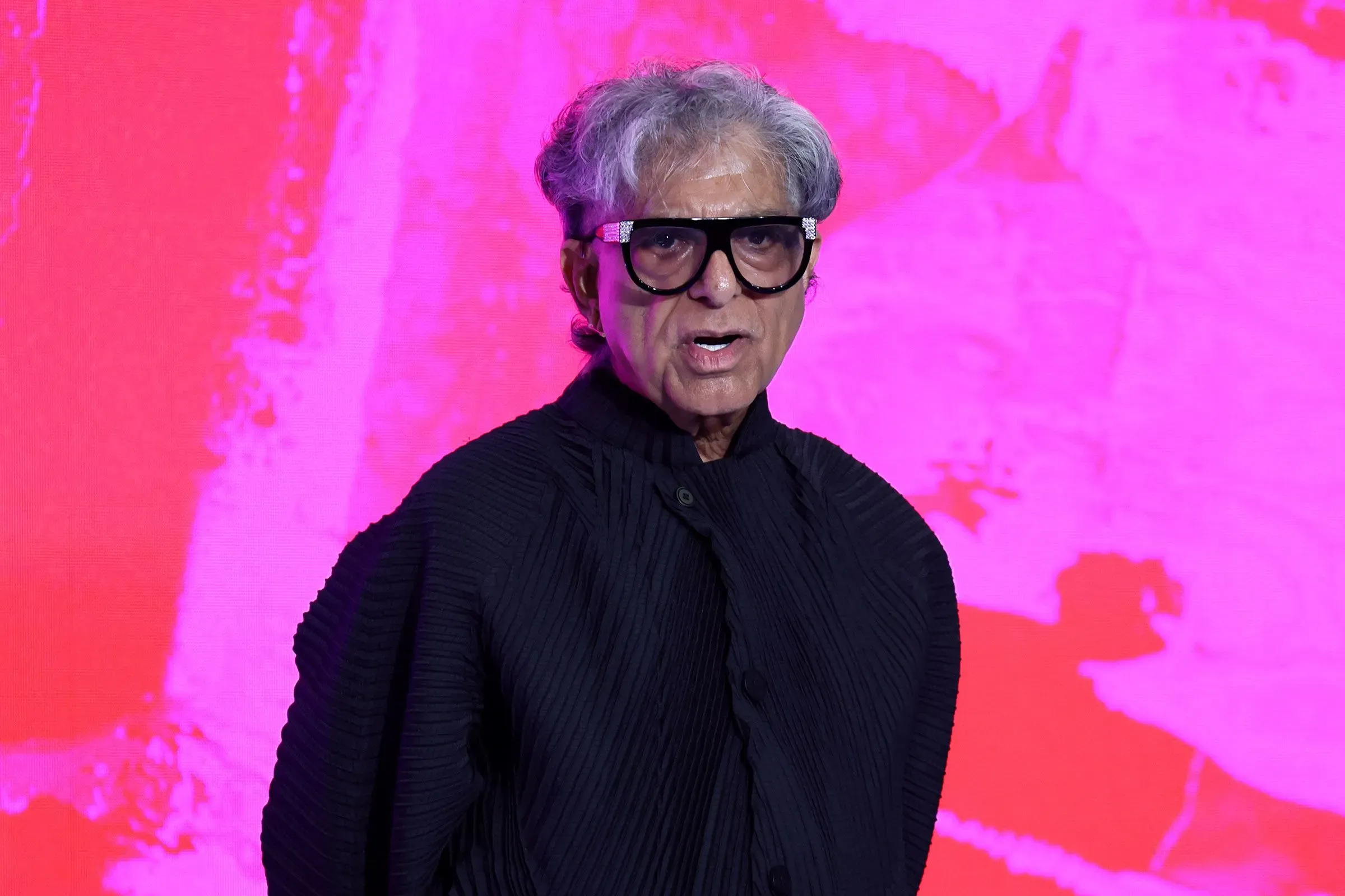 صورة لرجل مسن يرتدي نظارات وقميص أسود، يتحدث أمام خلفية ملونة وردية. تعكس الصورة أجواء حدث أو مؤتمر.