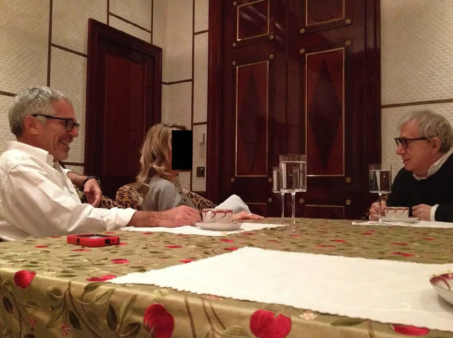 صورة تظهر ثلاثة أشخاص يجلسون حول طاولة مغطاة بقماش مزخرف، يتبادلون الحديث في بيئة داخلية فاخرة.