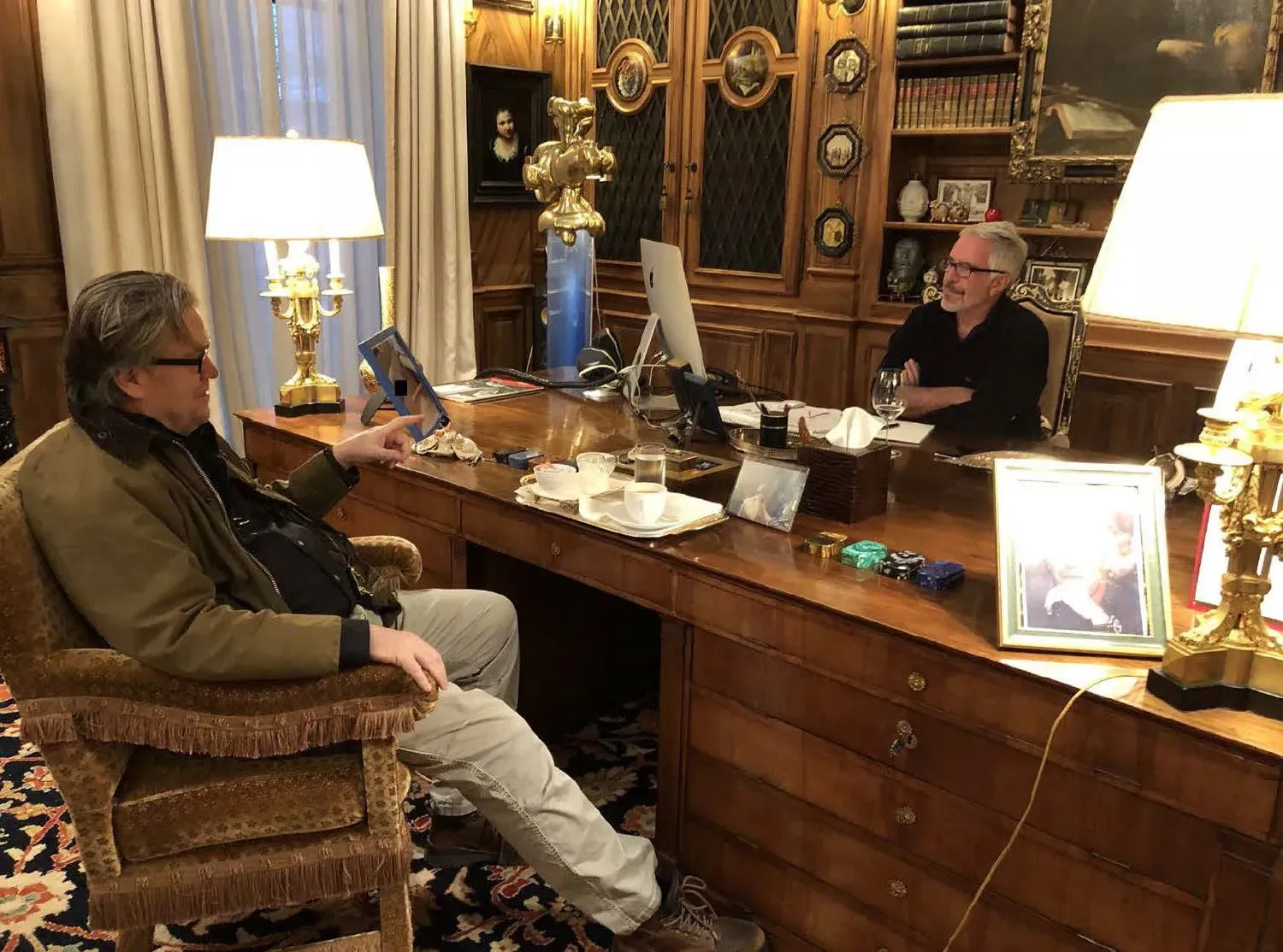 لقطة تظهر رجلين يجلسان في مكتب خشبي أنيق، حيث يتحدث أحدهما بينما يحمل الآخر كتابًا. الأجواء تعكس مناقشة غير رسمية.