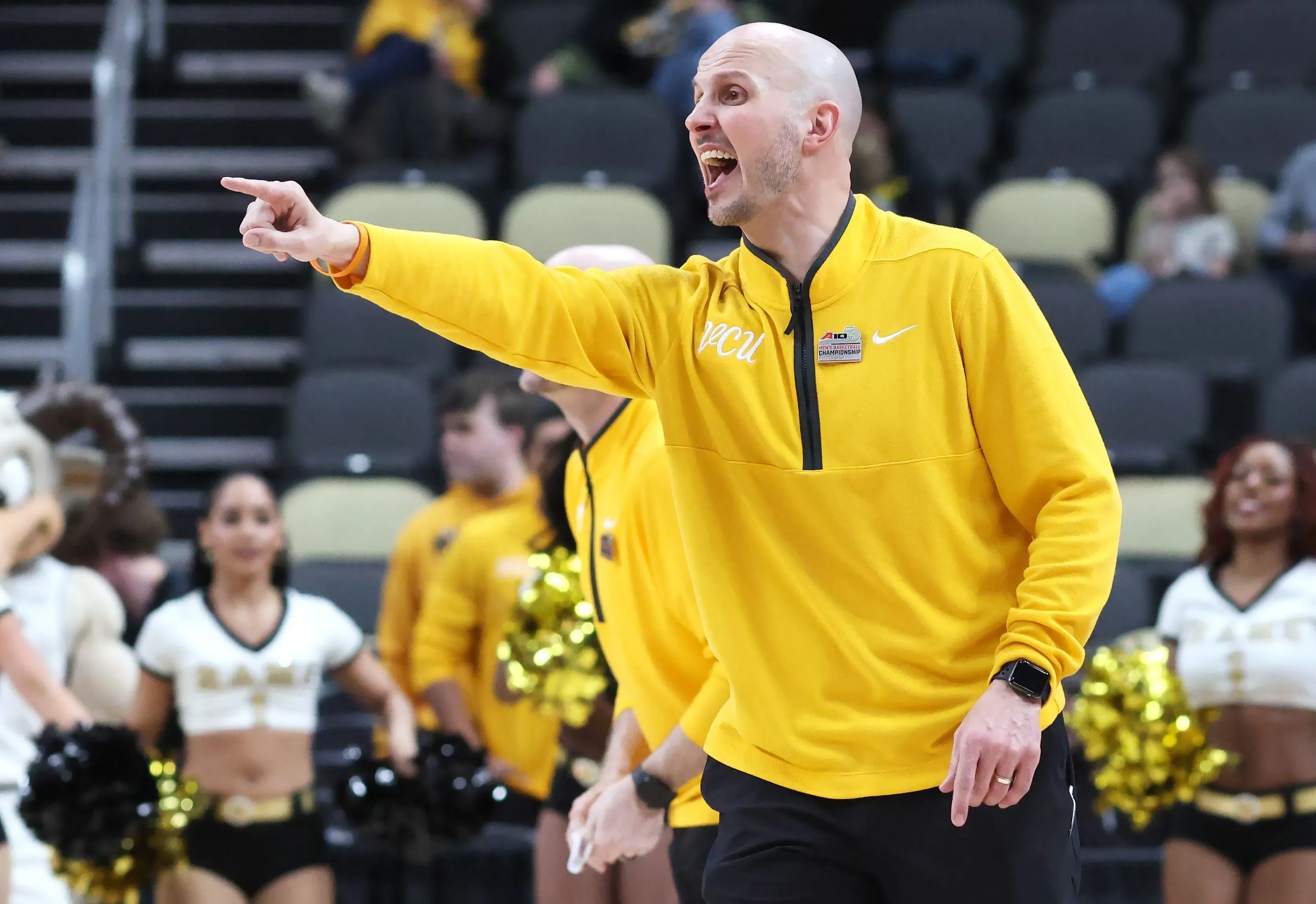 مدرب كرة سلة يرتدي زيًا أصفر ويشير بحماس أثناء توجيه فريقه في مباراة، مع وجود مشجعين خلفه في المدرجات.