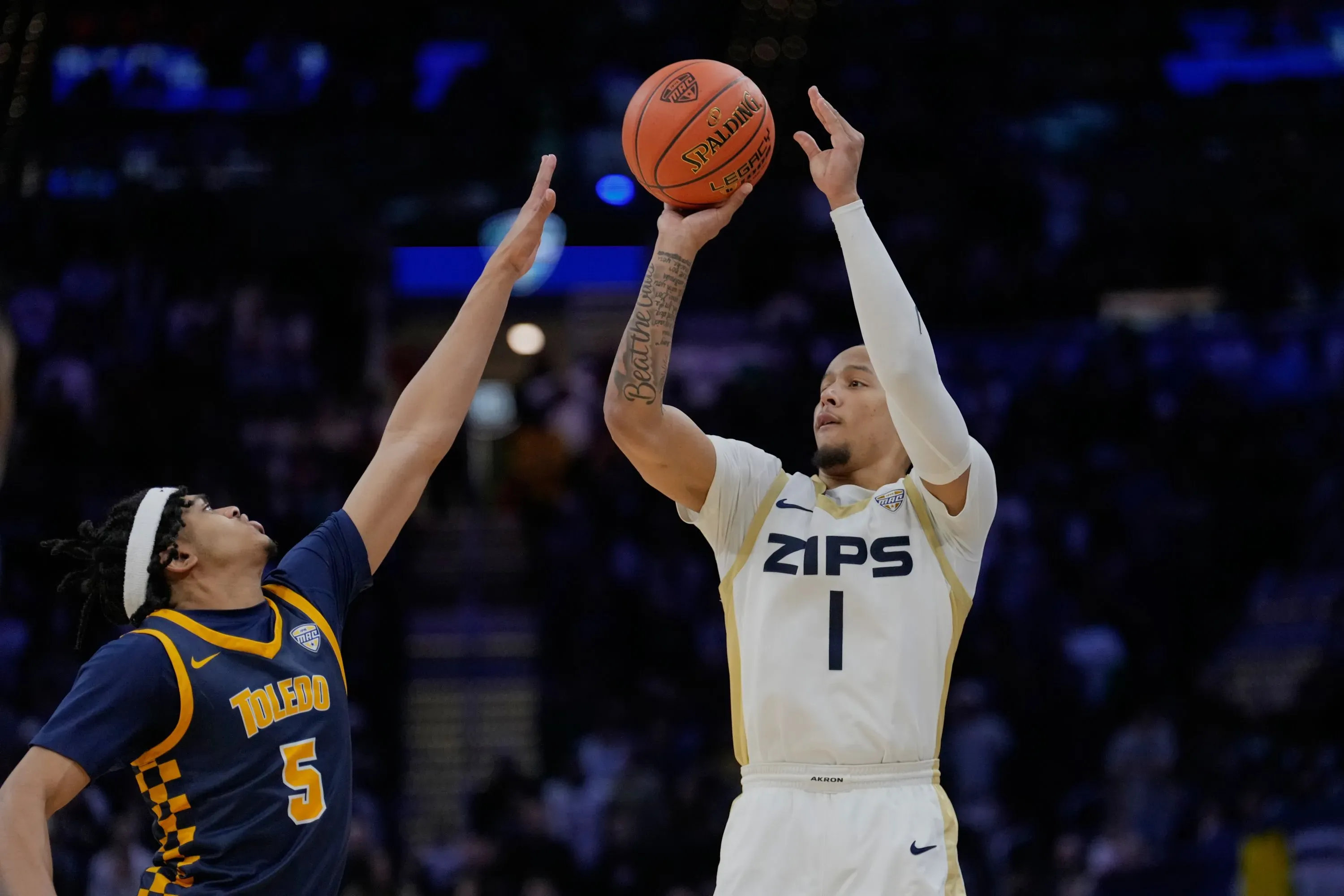 لاعب كرة سلة من فريق زيبس يسجل رمية ثلاثية بينما يحاول لاعب من فريق توليدو الدفاع عنه، في أجواء تنافسية حماسية.