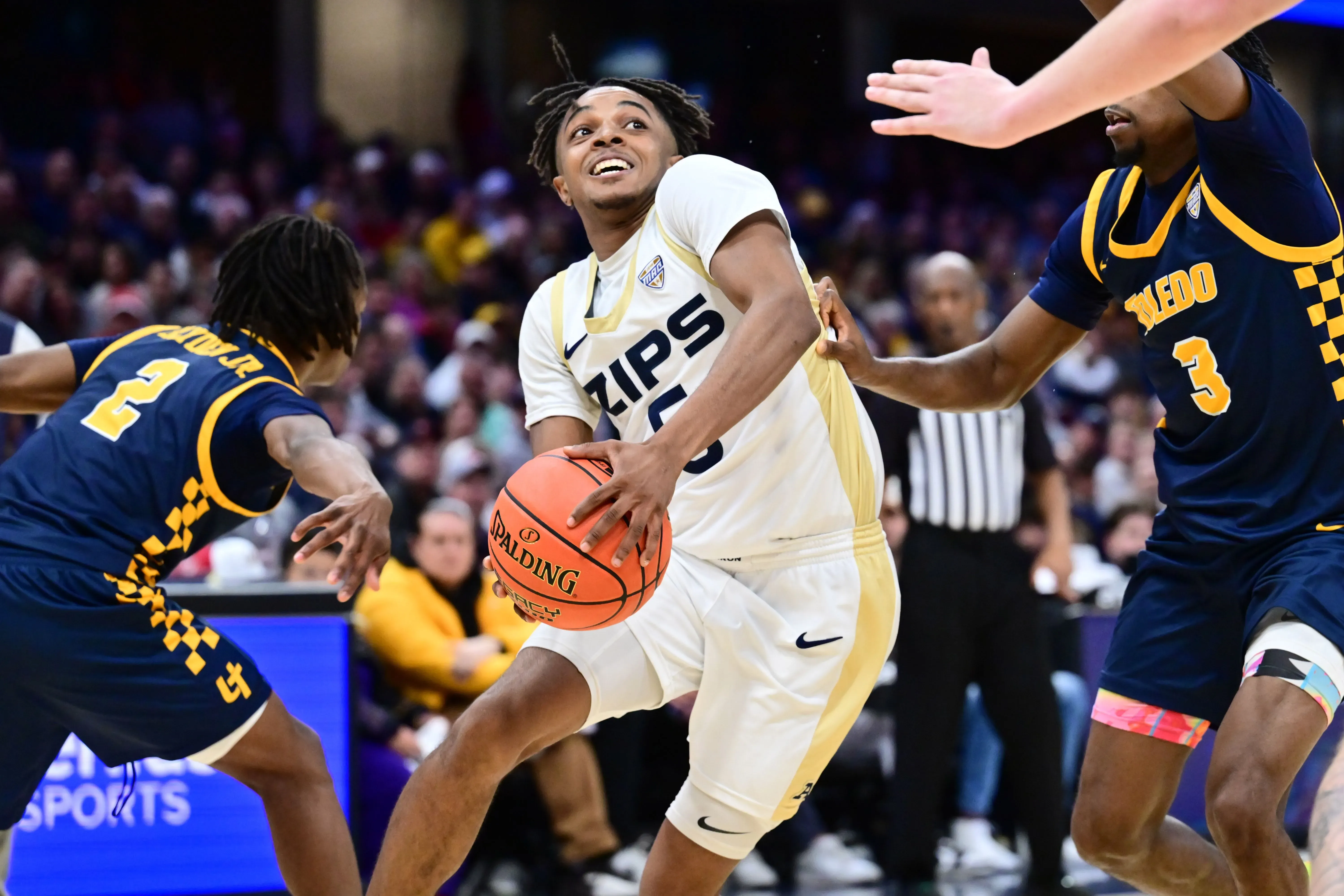 لاعب كرة سلة يرتدي زي فريق زيبس، يحاول المراوغة بينما يتعرض لضغط من لاعبين آخرين في مباراة حماسية.