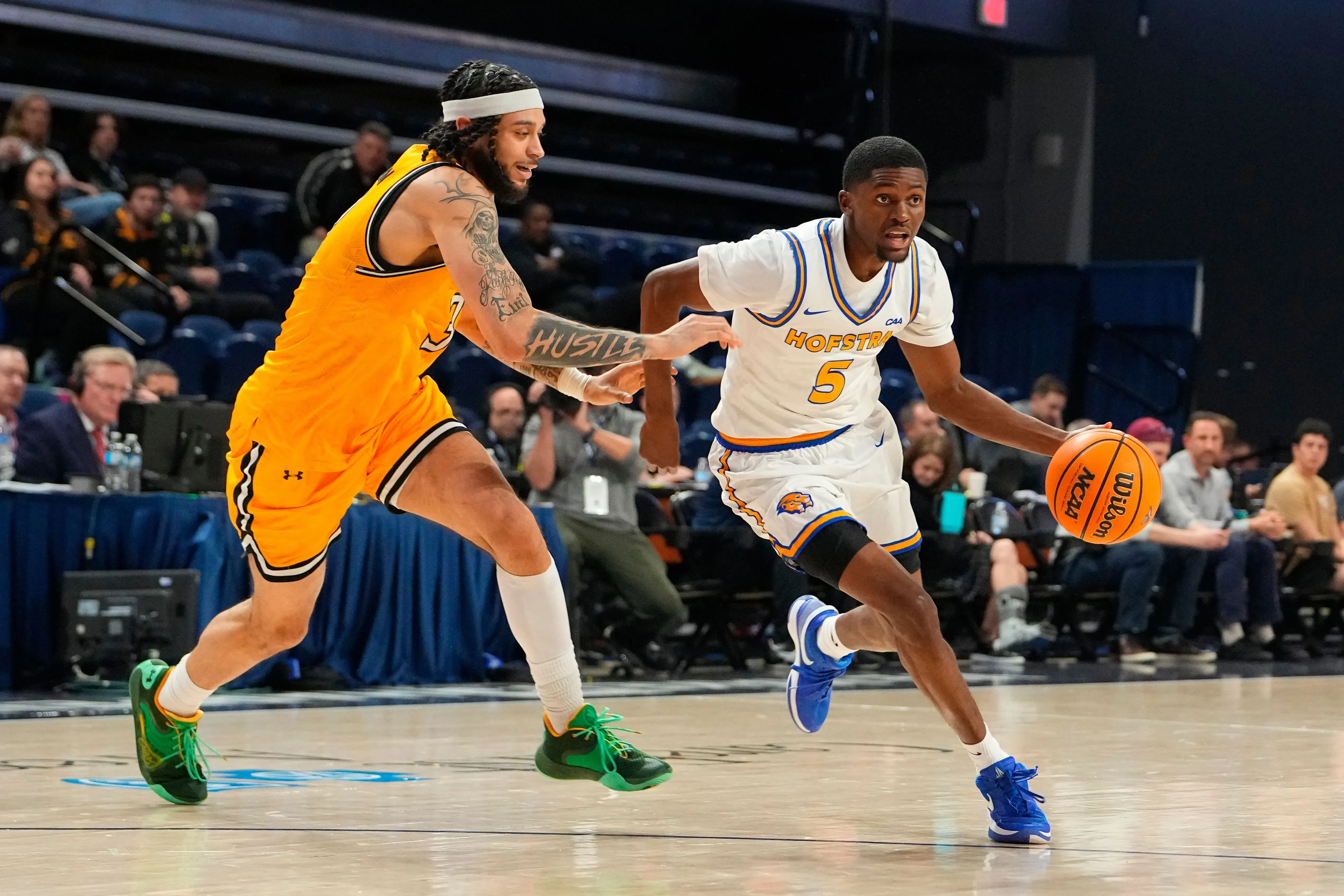 لاعب كرة سلة يرتدي زي هوفسترا الأبيض يحمل الكرة، بينما يتعقب لاعب آخر يرتدي زي أصفر. المباراة تجري في صالة رياضية مزدحمة.