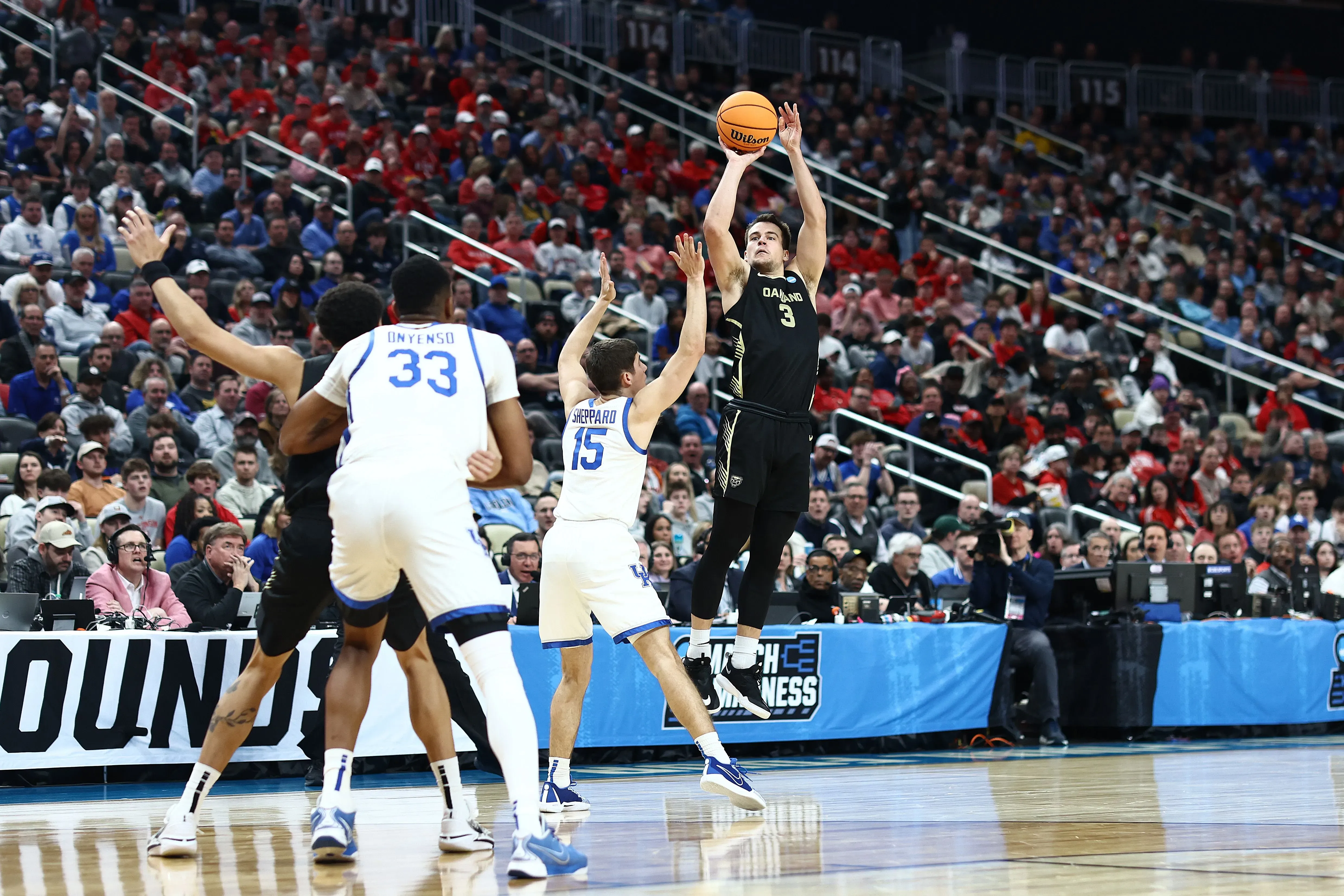 لاعب كرة سلة يرتدي زي فريقه الأسود، يقفز لتسديد كرة سلة، بينما يحاول لاعبون من الفريق المنافس اعتراضه في مباراة NCAA.