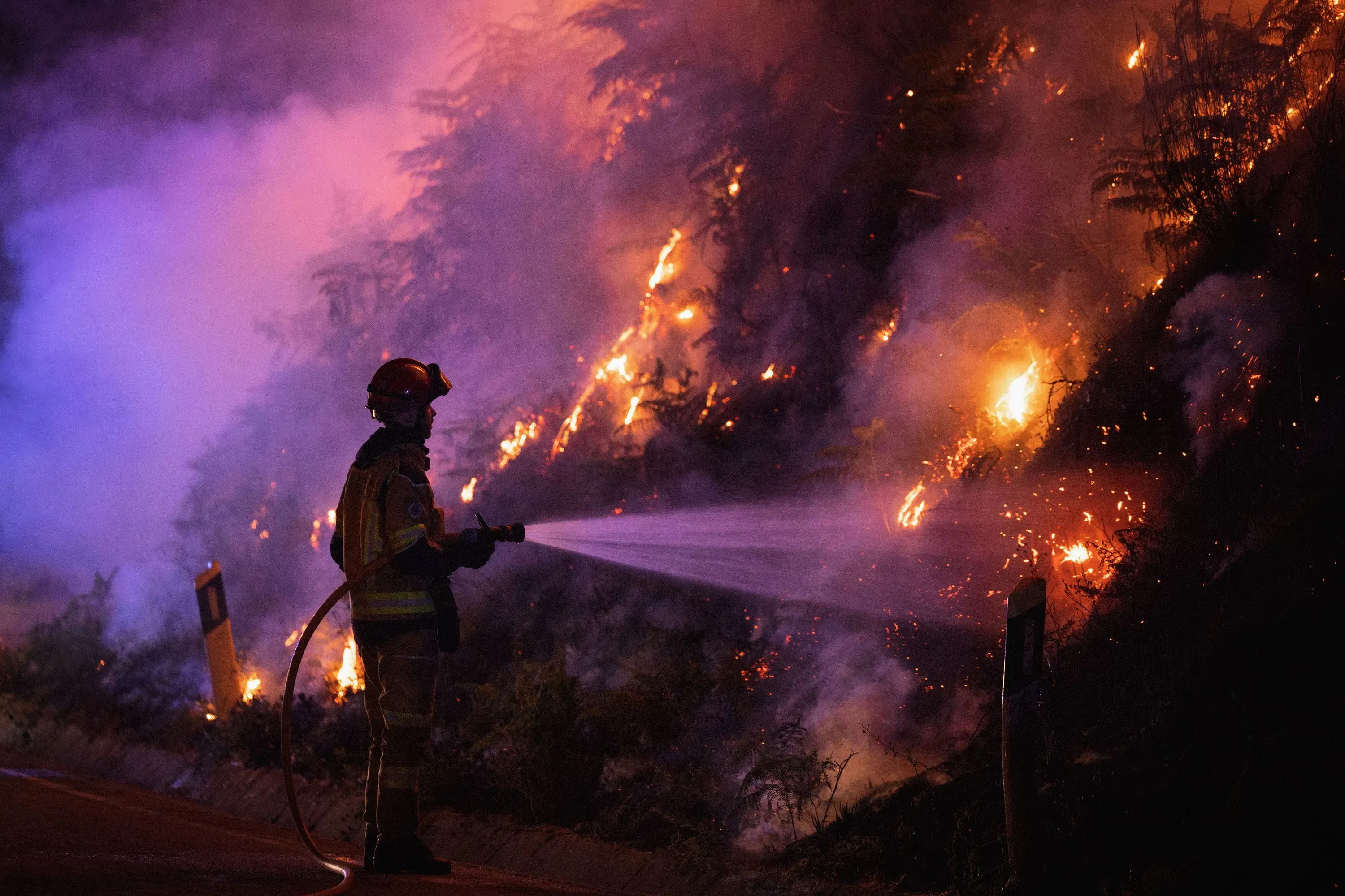 رجال الإطفاء يكافحون حرائق الغابات في منطقة مشتعلة، حيث تتصاعد الدخان واللهب في الليل، مما يعكس تأثيرات موجات الحر المتزايدة.