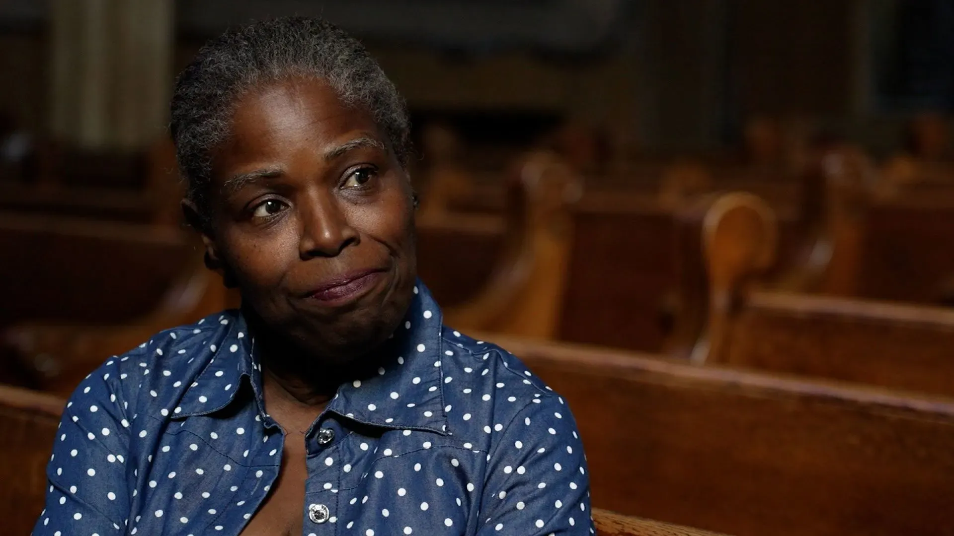 امرأة ذات بشرة داكنة ترتدي قميصًا مزخرفًا بنقاط بيضاء، تجلس في كنيسة، تعبر عن قلقها بشأن العنف المسلح في حيها.