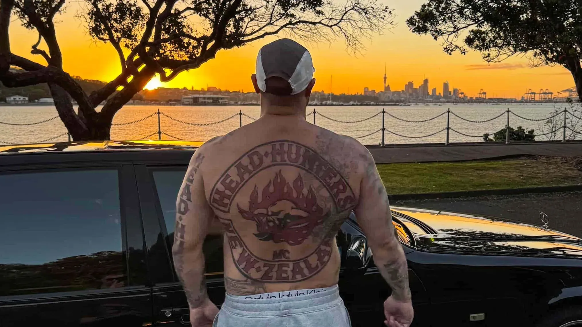 رجل ذو وشم كبير على ظهره يحمل اسم "صائدو الرؤوس" مع جمجمة وألسنة لهب، يقف أمام منظر غروب الشمس في نيوزيلندا.