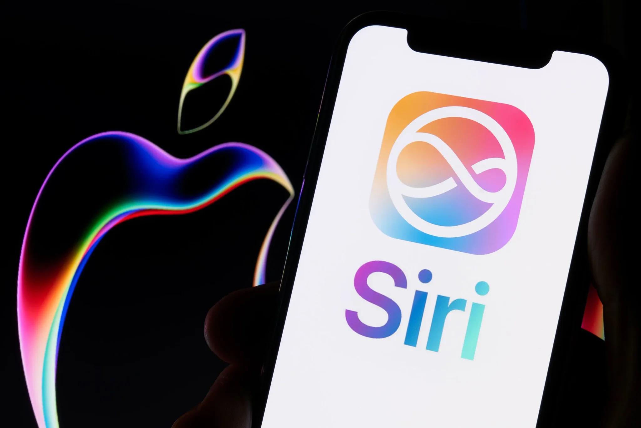 شعار Siri الملون على شاشة هاتف iPhone مع خلفية شعار Apple بألوان متدرجة، يعكس تحديث iOS 17 وتحول Siri إلى مساعد ذكي متقدم.