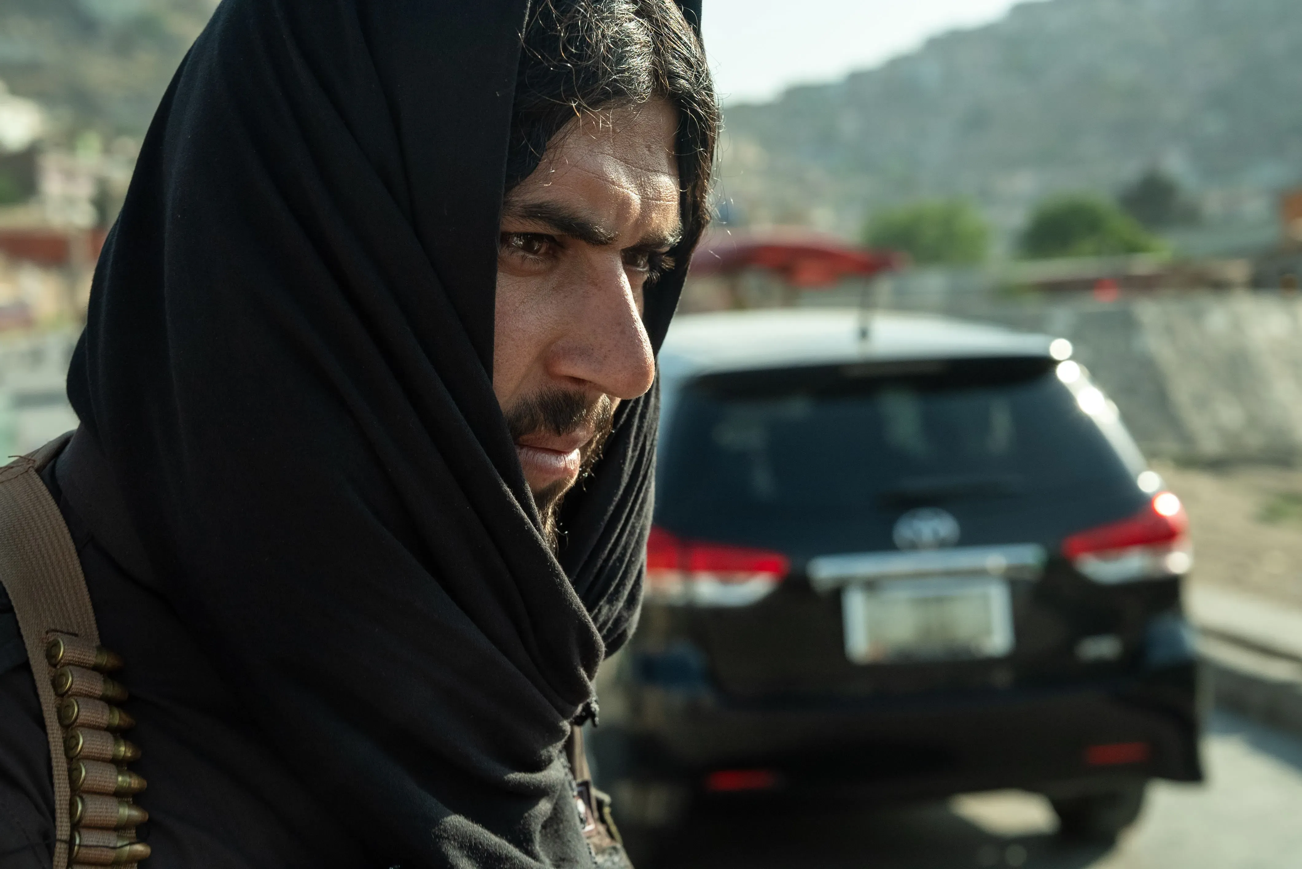 رجل يرتدي غطاء رأس أسود، يبدو جادًا، يقف في الشارع بالقرب من سيارة، مما يعكس الوضع الراهن في أفغانستان تحت حكم طالبان.