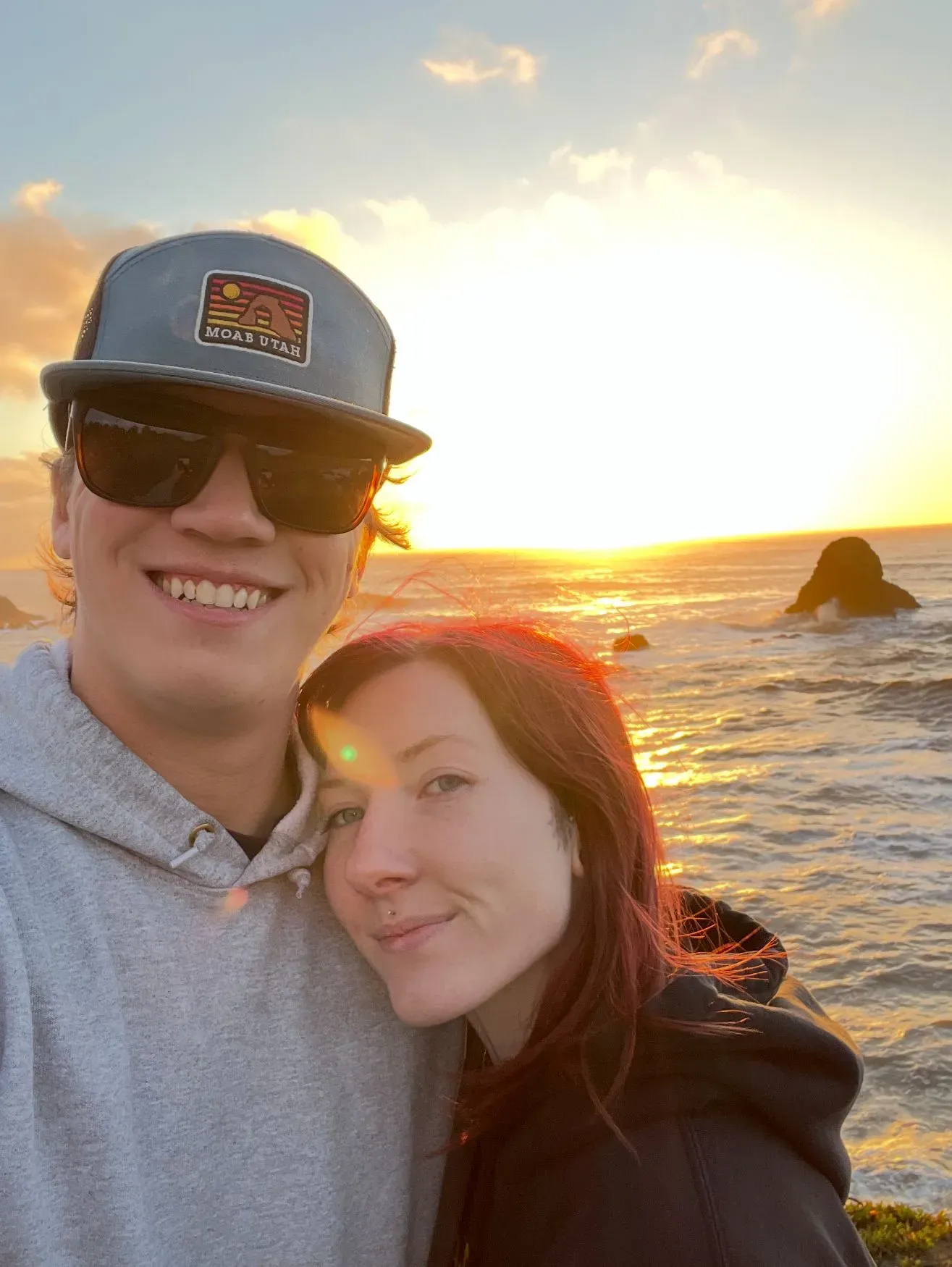 زوجان يتعانقان مبتسمين أمام غروب الشمس على الشاطئ، مع وجود صخور في الأفق، معبرين عن لحظة من السعادة وسط تحديات الحياة.