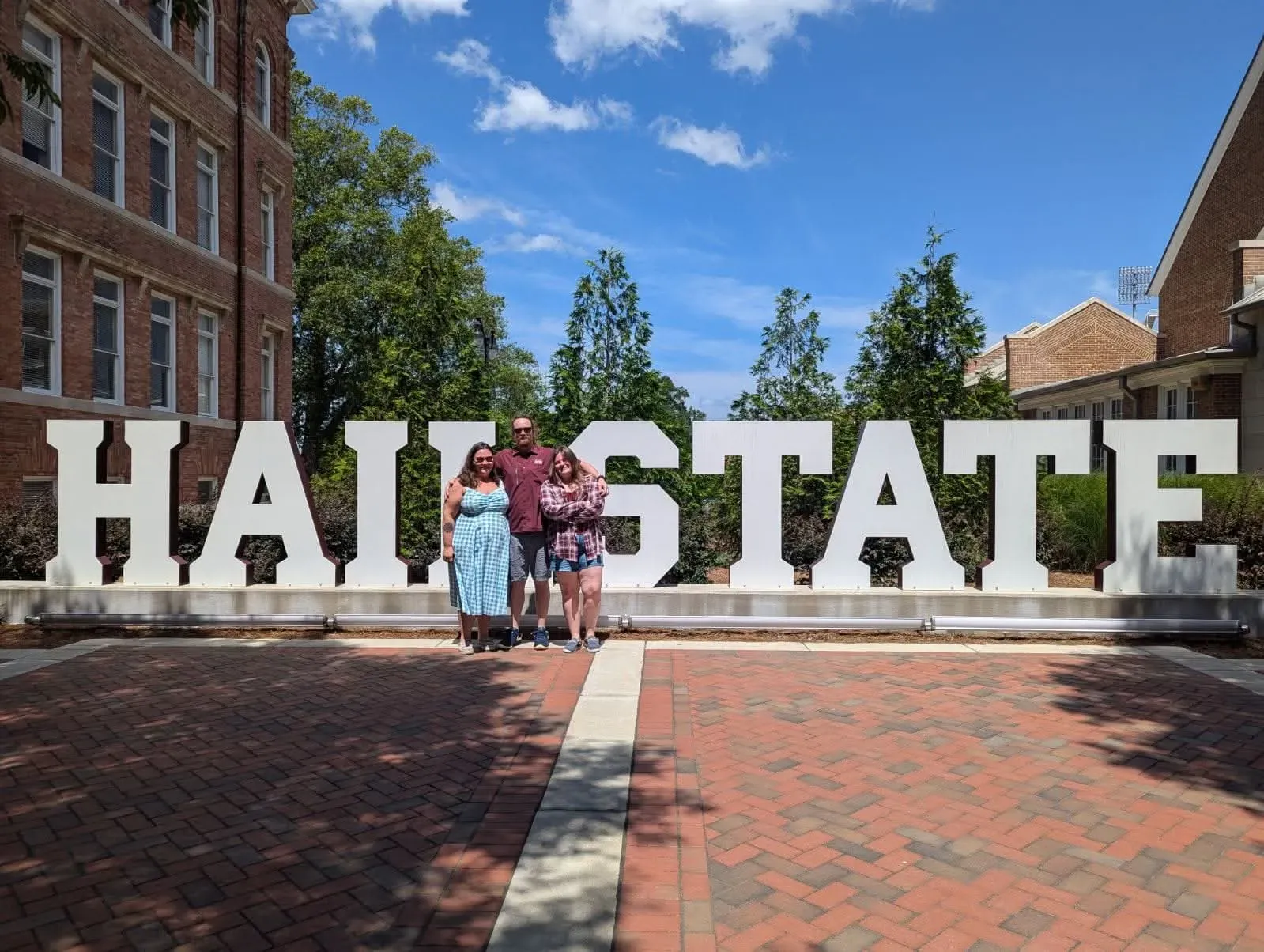 مجموعة من الأشخاص يقفون أمام لافتة كبيرة مكتوب عليها "HAIL STATE" في حرم جامعي، مما يعكس روح المجتمع الجامعي في كانساس.