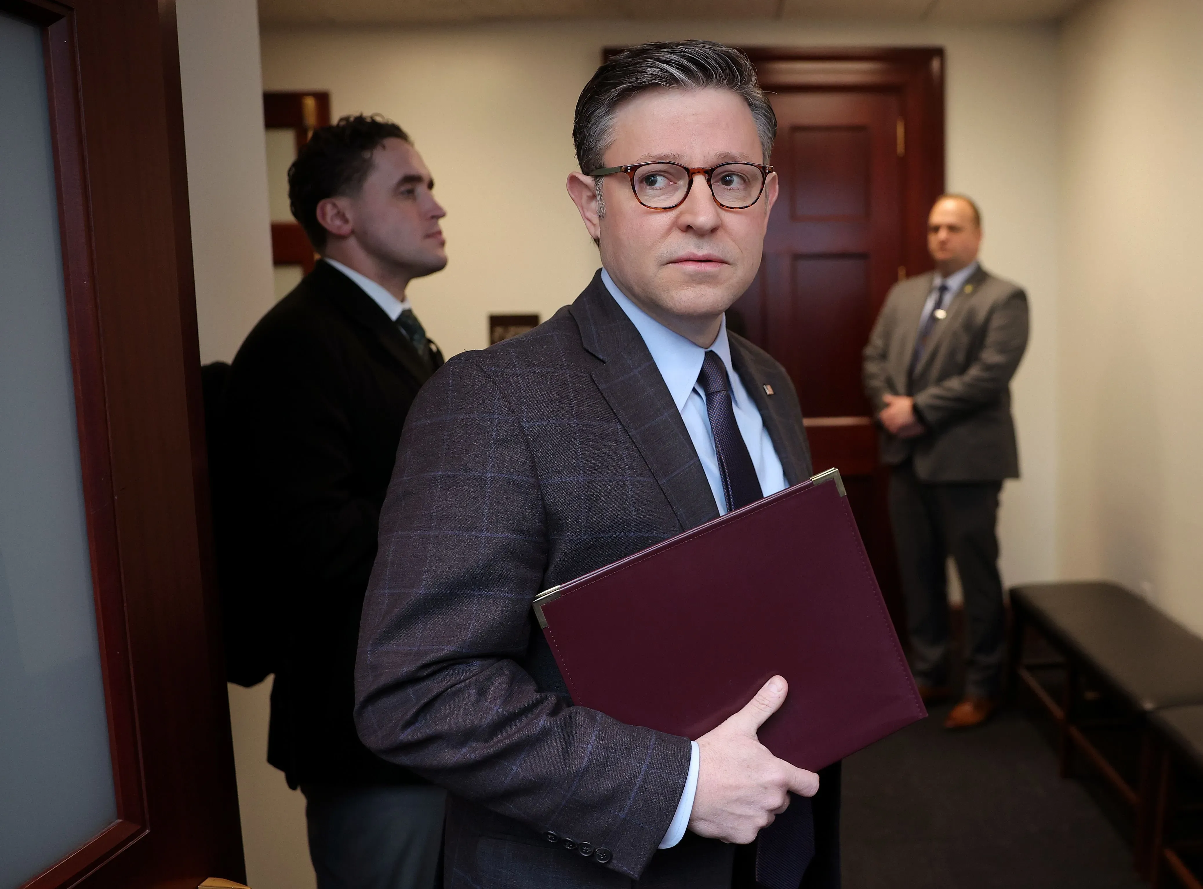 رجل يحمل ملفًا في ممر بمبنى حكومي، يبدو عليه القلق، بينما يقف رجلان آخران خلفه في انتظار. تعكس الصورة توتر المناقشات حول ميزانية الحكومة الفيدرالية.