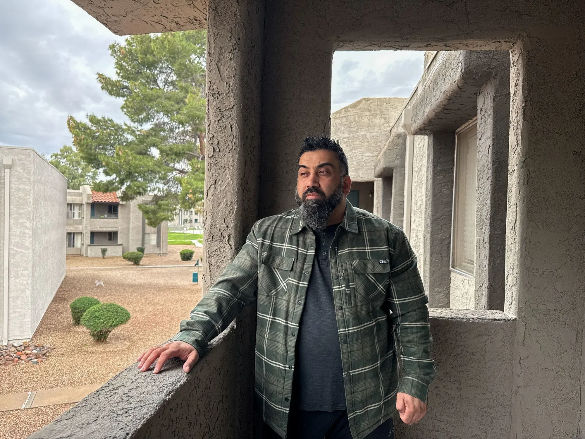 رجل يقف على شرفة شقته، ينظر بقلق إلى الخارج، في خلفية معمارية حديثة، مما يعكس مشاعر التوتر بشأن قضايا الهجرة.