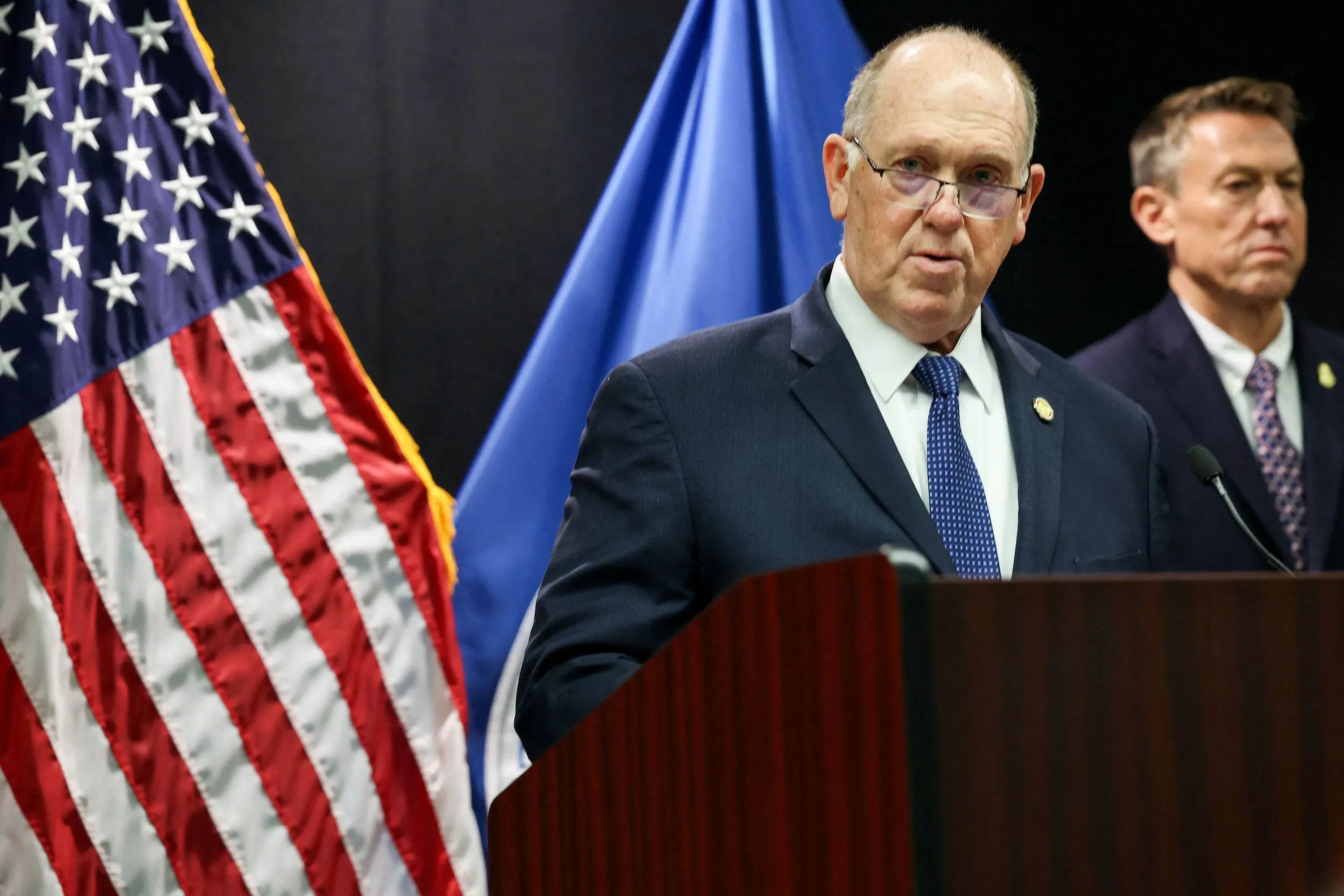 متحدث رسمي يقف أمام منصة خلال مؤتمر صحفي، مع العلم الأمريكي خلفه، يناقش قضايا الهجرة واللاجئين في الولايات المتحدة.