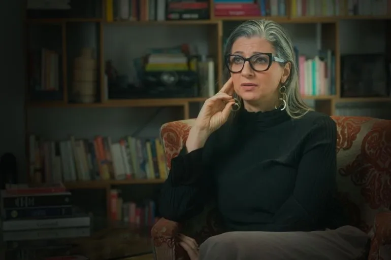 امرأة تجلس في غرفة مع كتب خلفها، تعبر عن مشاعرها حول قضايا الاعتداء الجنسي في السجون الإسرائيلية، في سياق تحقيق حول حقوق الإنسان.