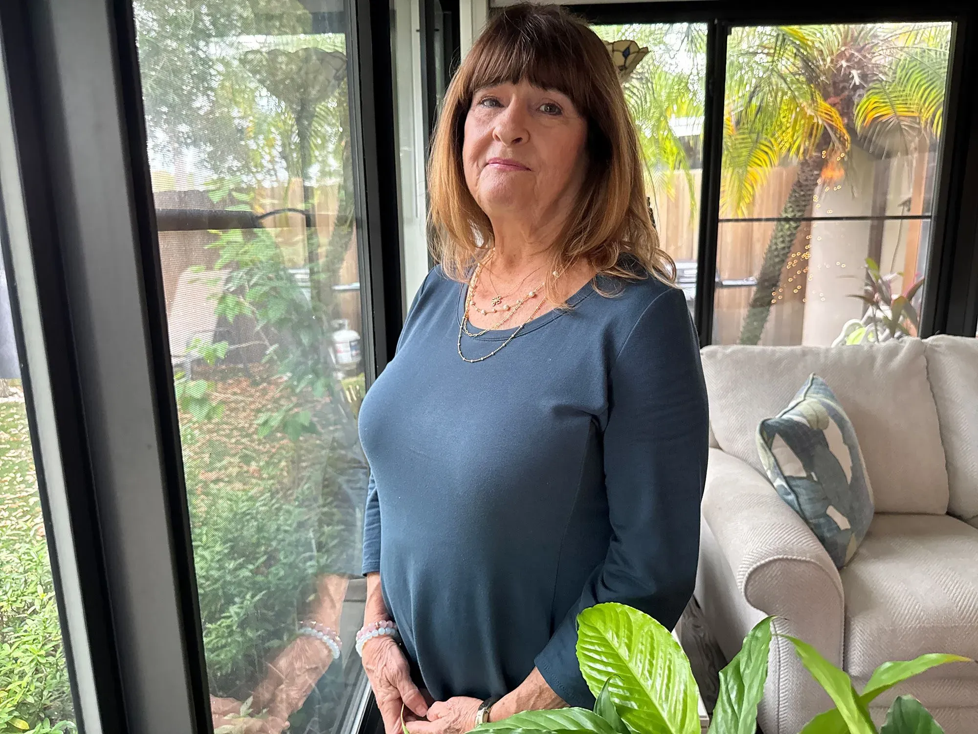 امرأة مسنّة تقف بجانب نافذة في منزلها، تعبر عن مشاعرها بعد فقدان زوجها، مع نباتات خضراء في الخلفية.