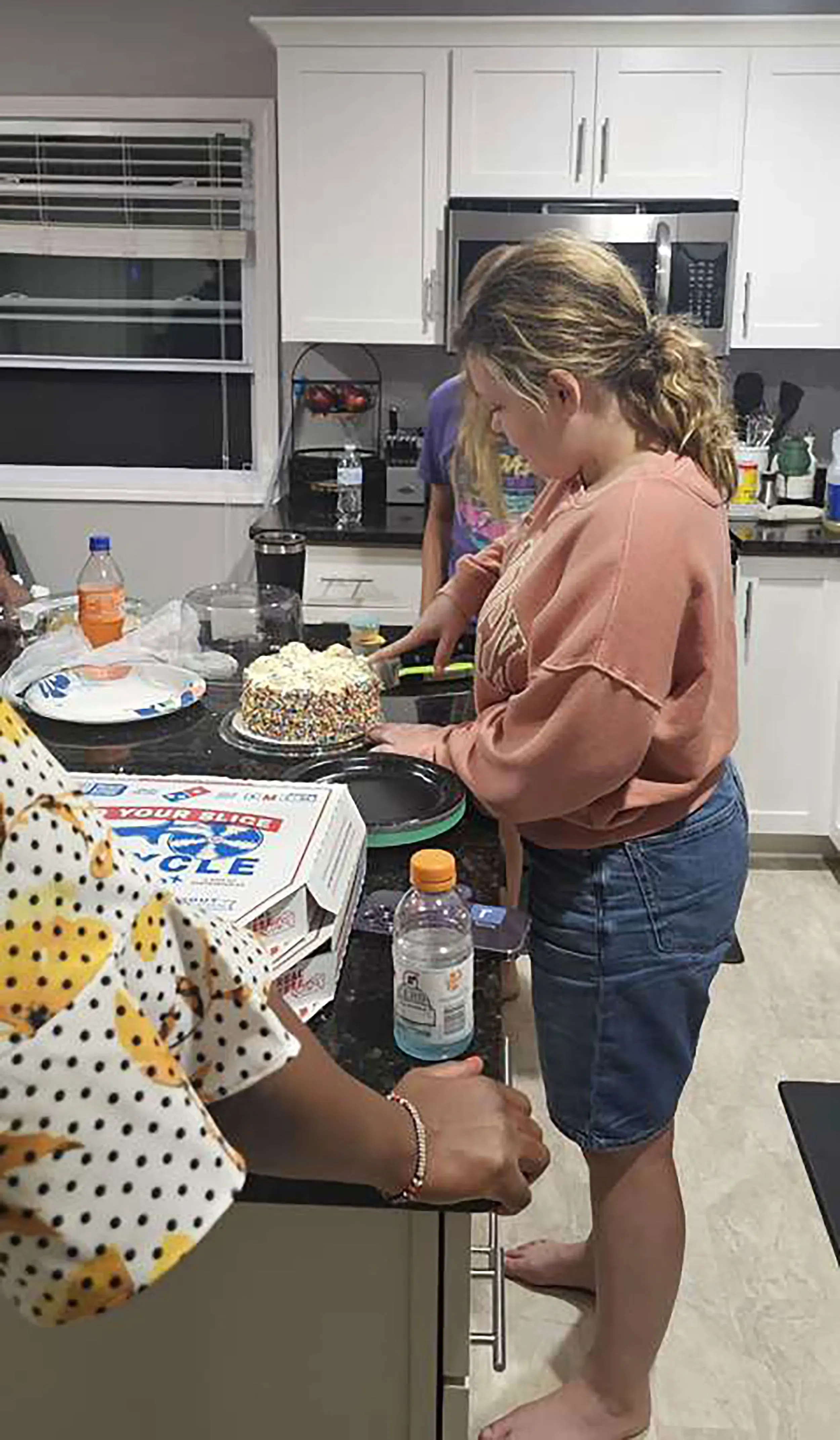 فتاة شابة تقطع كعكة ملونة في مطبخ حديث، بينما تظهر علب البيتزا والمشروبات في الخلفية، تعكس لحظة احتفال عائلي.