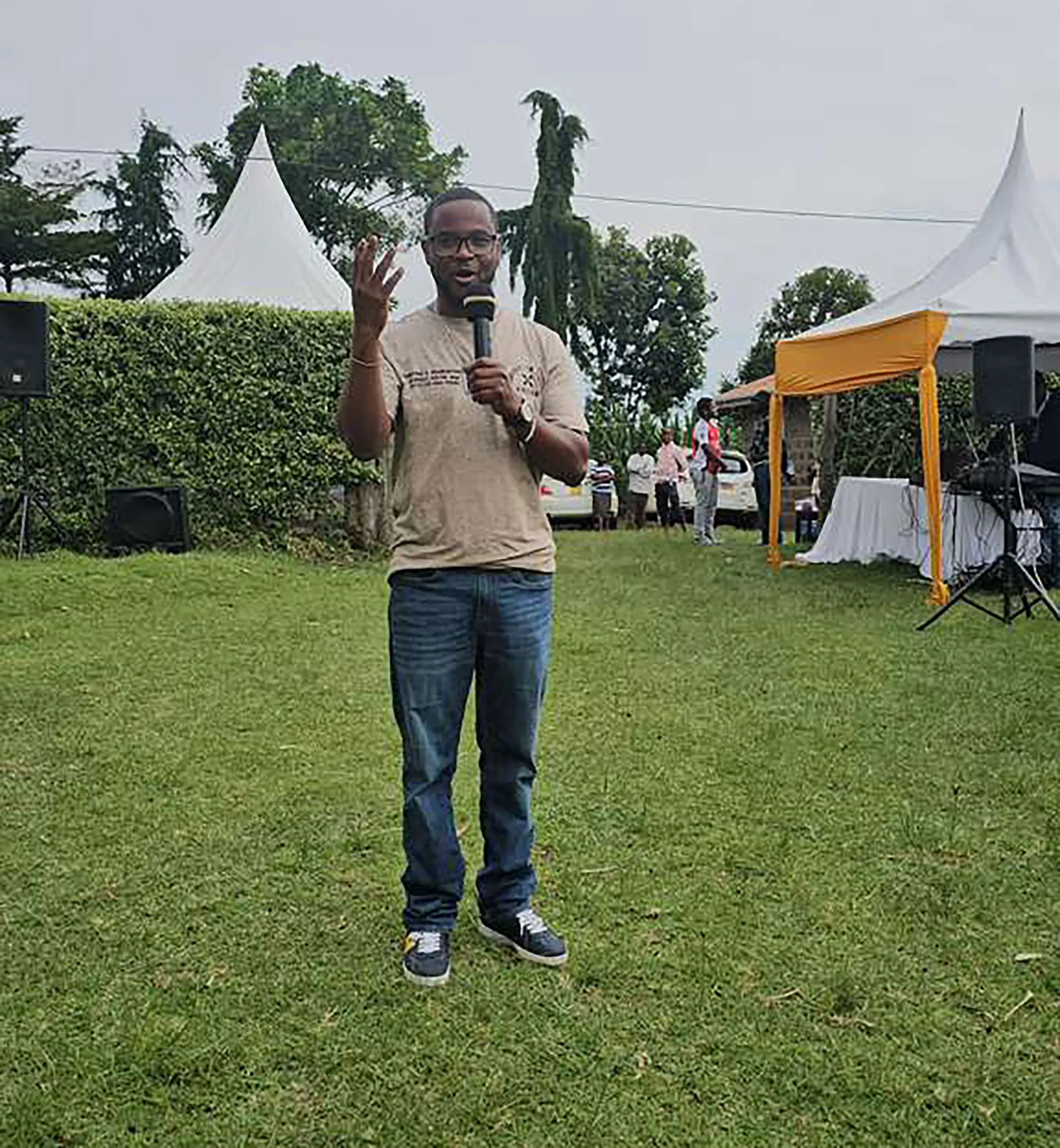 كانغيث يتحدث في حدث خارجي، مرتديًا قميصًا بنيًا وسروالًا جينز، بينما يرفع يده للتعبير عن نقطة مهمة. خلفه، تظهر خيمة بيضاء وأشخاص يتجمعون في حديقة خضراء.