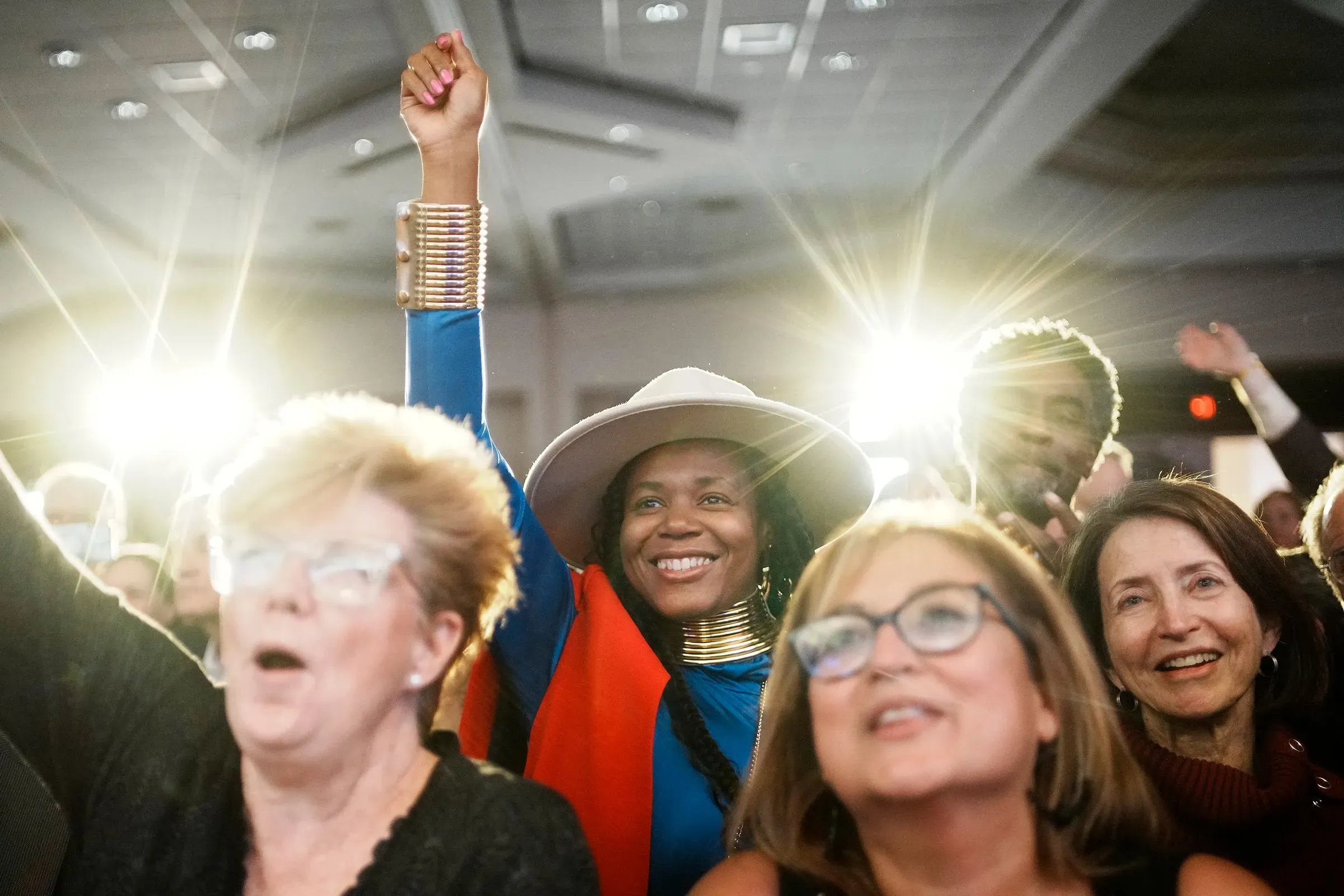 نساء ورجال في حدث سياسي، يظهرون الحماس والتفاعل، مع رفع أحدهم يده في الهواء. الأضواء تسلط عليهم، مما يبرز أجواء الاحتفال.