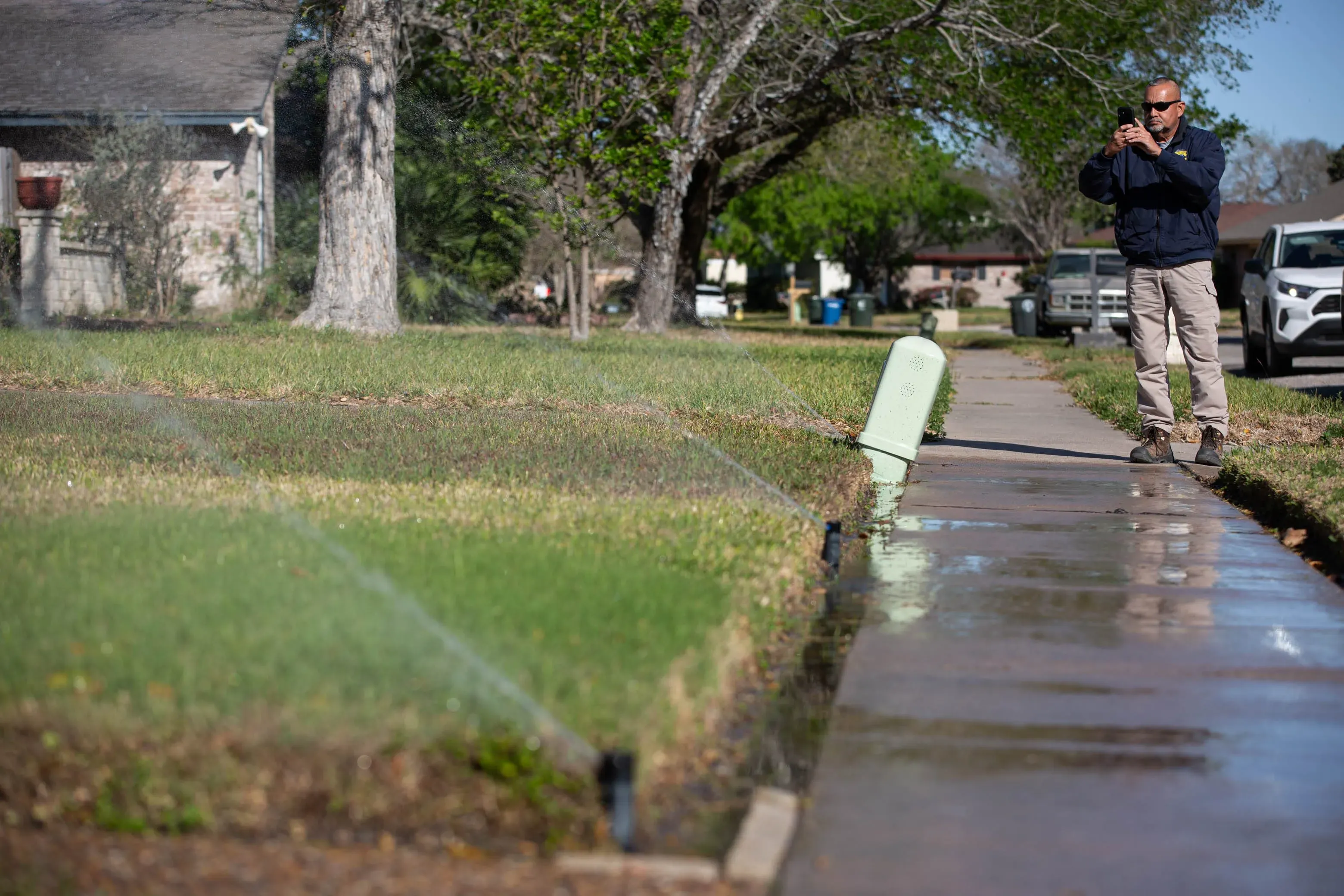 رجل يقف على الرصيف بينما يلتقط صورة لحديقة تسقى بالمياه، مع وجود أشجار ومنازل في الخلفية، في سياق أزمة المياه في كوربوس كريستي.