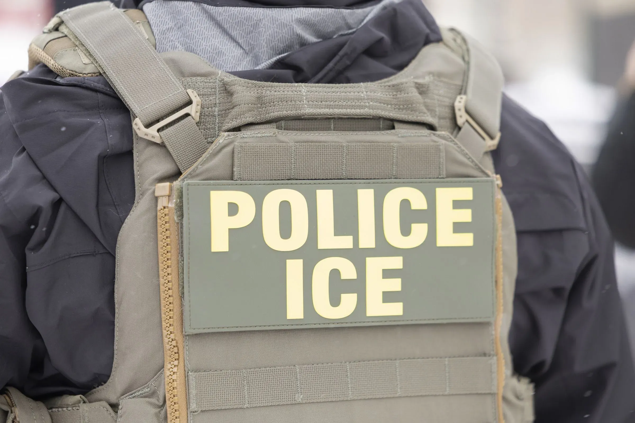صورة لظهر ضابط يرتدي سترة واقية تحمل كلمة "شرطة" و"ICE"، مما يشير إلى وجود عمليات إنفاذ قوانين الهجرة.