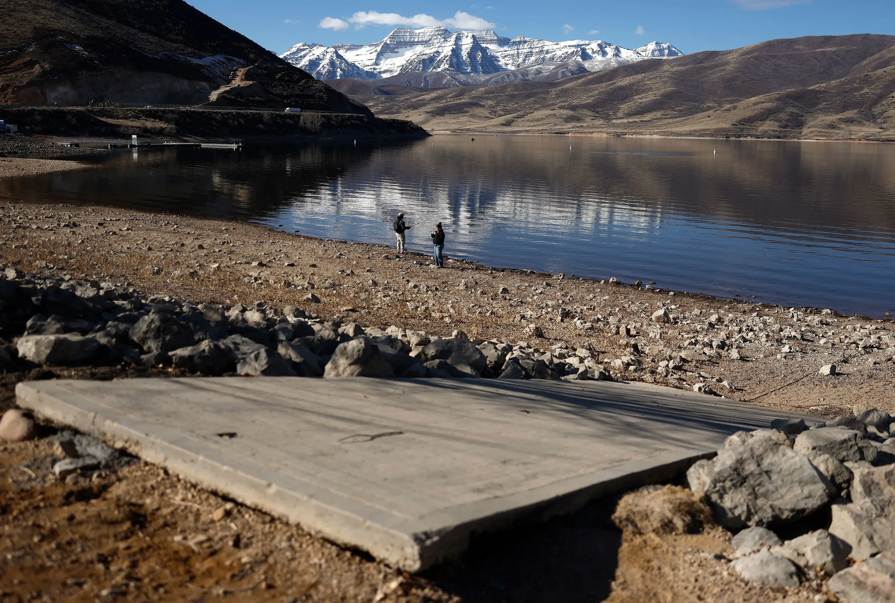 تظهر الصورة شخصين يسيران على ضفاف بحيرة جافة في منطقة جبلية، مع خلفية من الجبال المغطاة بالثلوج، مما يعكس تأثيرات تغير المناخ.