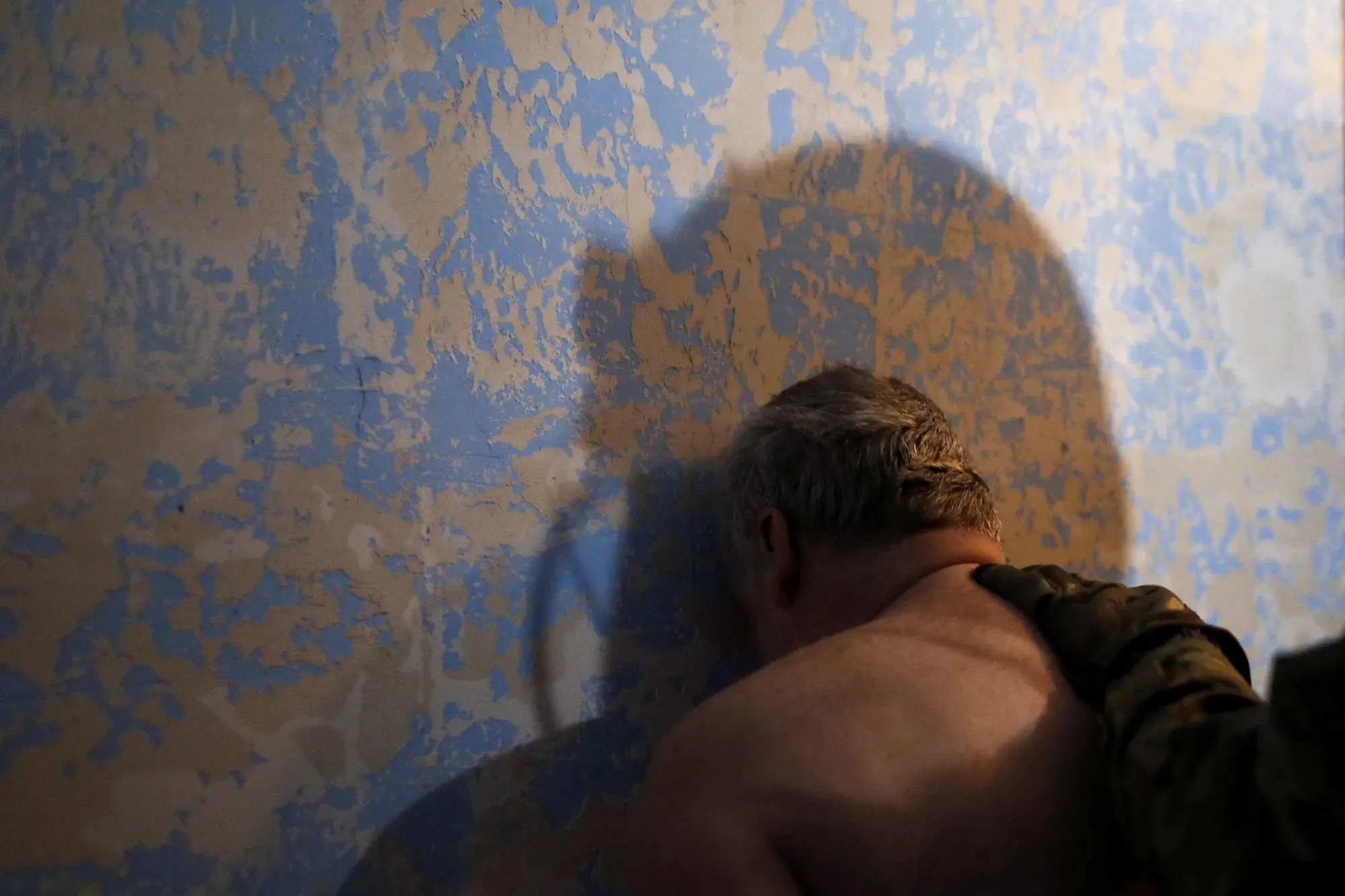 رجل يتكئ على حائط متصدع بلون أزرق، بينما يظهر ظل شخص آخر خلفه، مما يعكس حالة توتر أو استجواب في سياق الحرب.