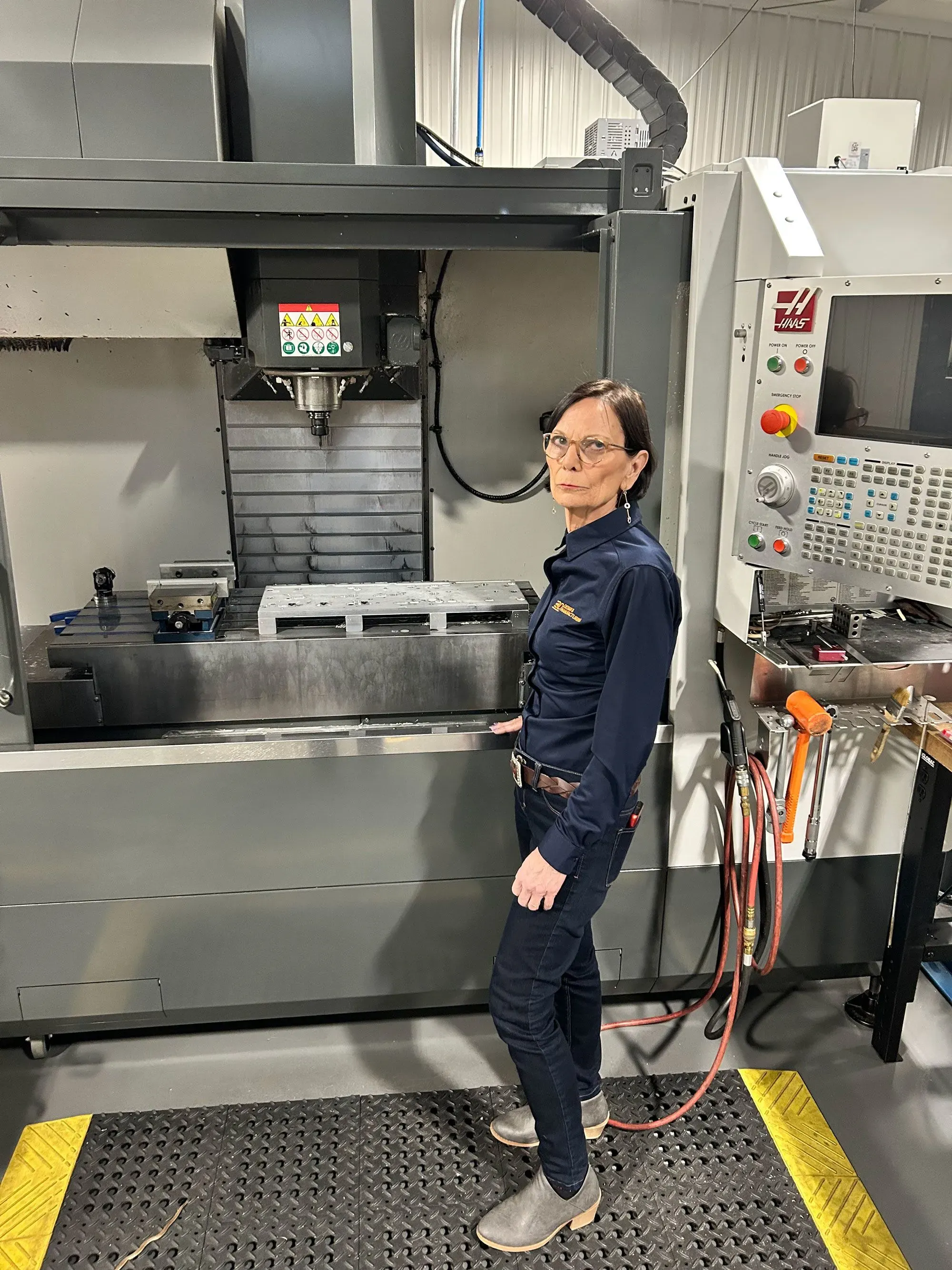 امرأة تقف بجانب آلة تصنيع حديثة في مصنع، ترتدي ملابس عمل، وتظهر ملامح التركيز على وجهها.