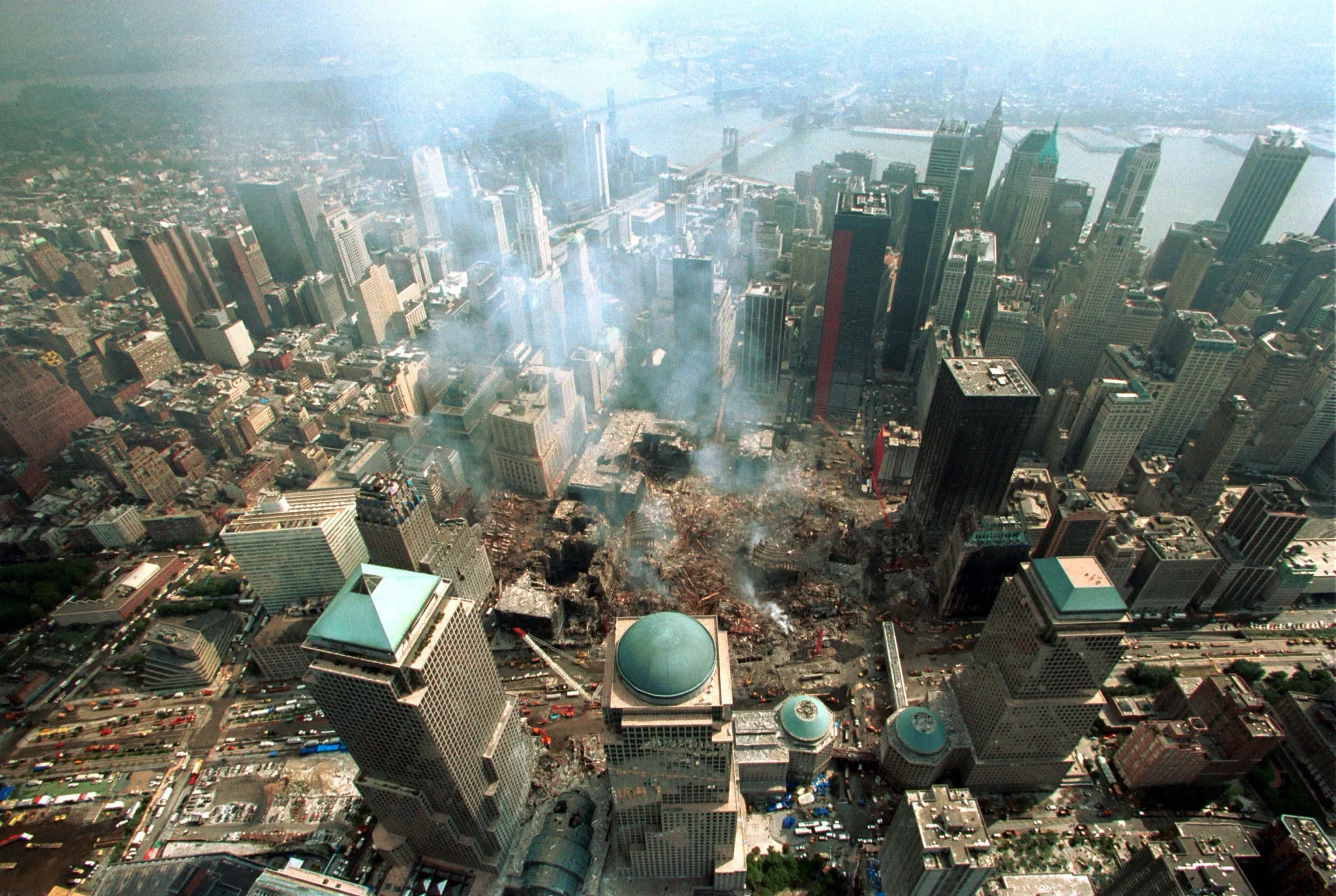 دخان وركام موقع هجمات 11 سبتمبر 2001 في وسط مدينة نيويورك، مع مبانٍ مرتفعة تحيط بالمكان، يعكس آثار الكارثة والهواء السام الذي تسبب بأمراض وسرطان للمنقذين والسكان.