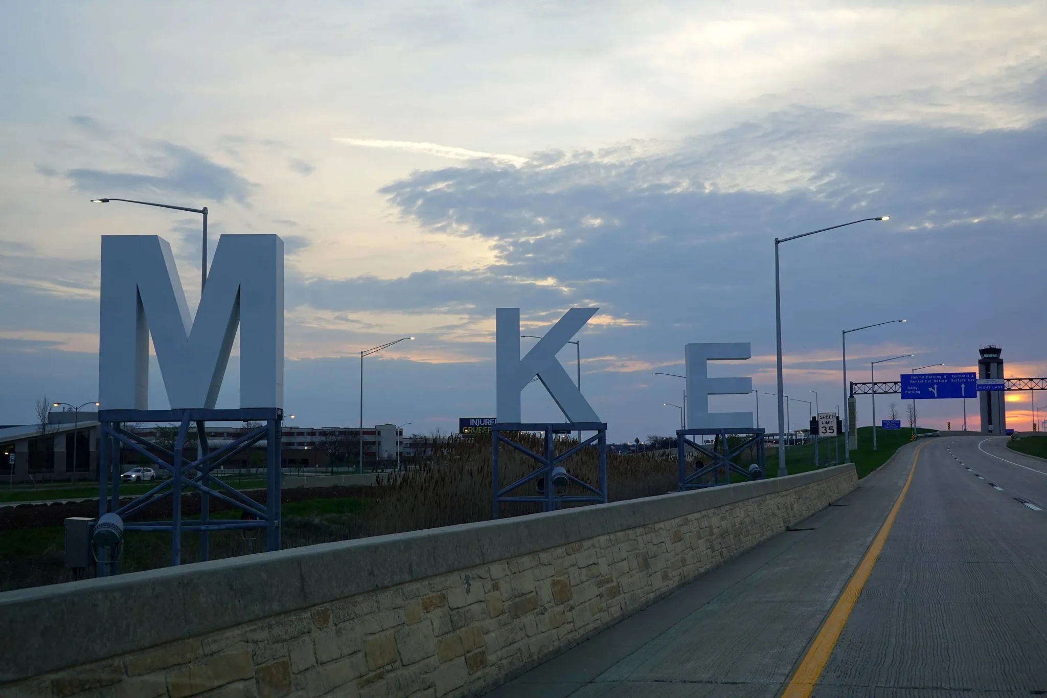 لافتات كبيرة بحروف M K E على طريق سريع قرب مطار Milwaukee في غروب الشمس، تعكس مكان وقوع حادثة تهديد على متن طائرة United Airlines.
