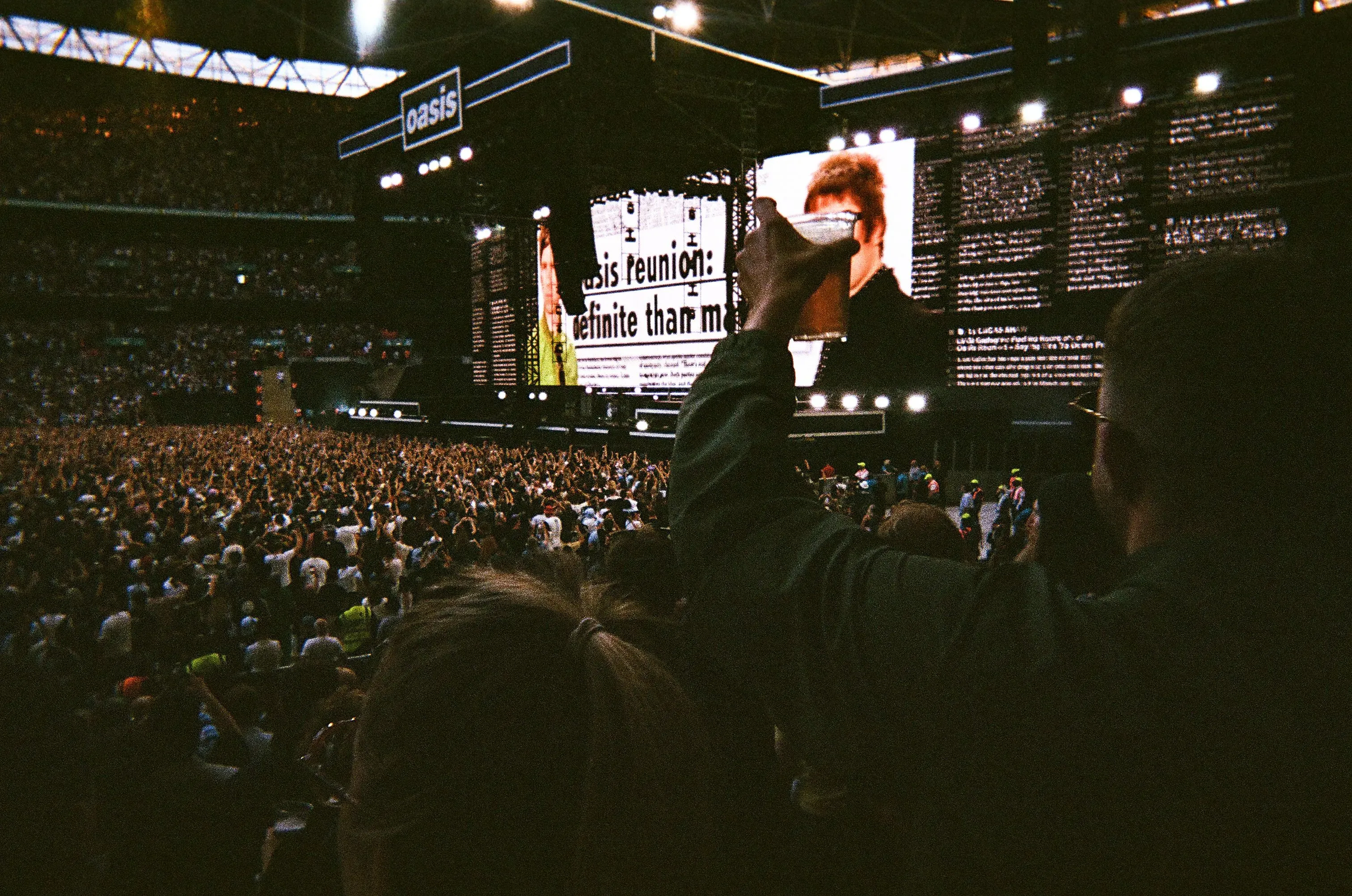 حشود كبيرة من المعجبين في ملعب ويمبلي خلال حفل فرقة Oasis، مع عرض كلمات الأغاني على الشاشة الخلفية.