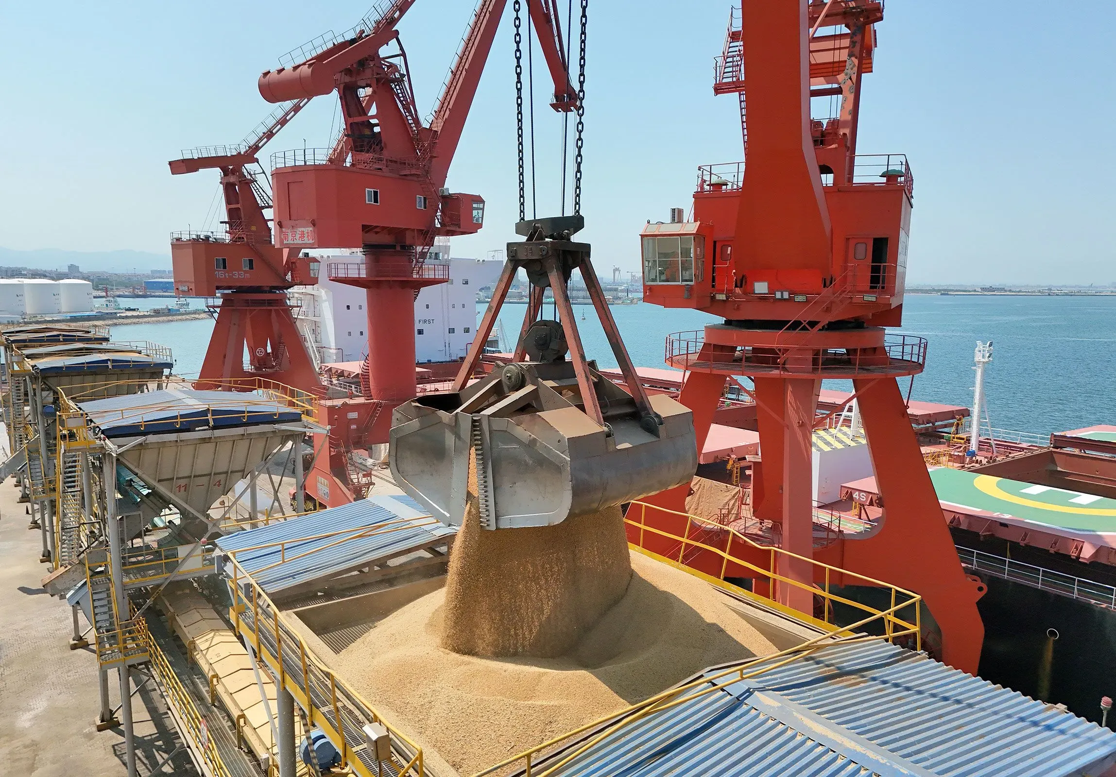 رافعة تحميل ضخمة تقوم بنقل كميات كبيرة من فول الصويا في ميناء، مع وجود معدات تحميل حمراء في الخلفية.