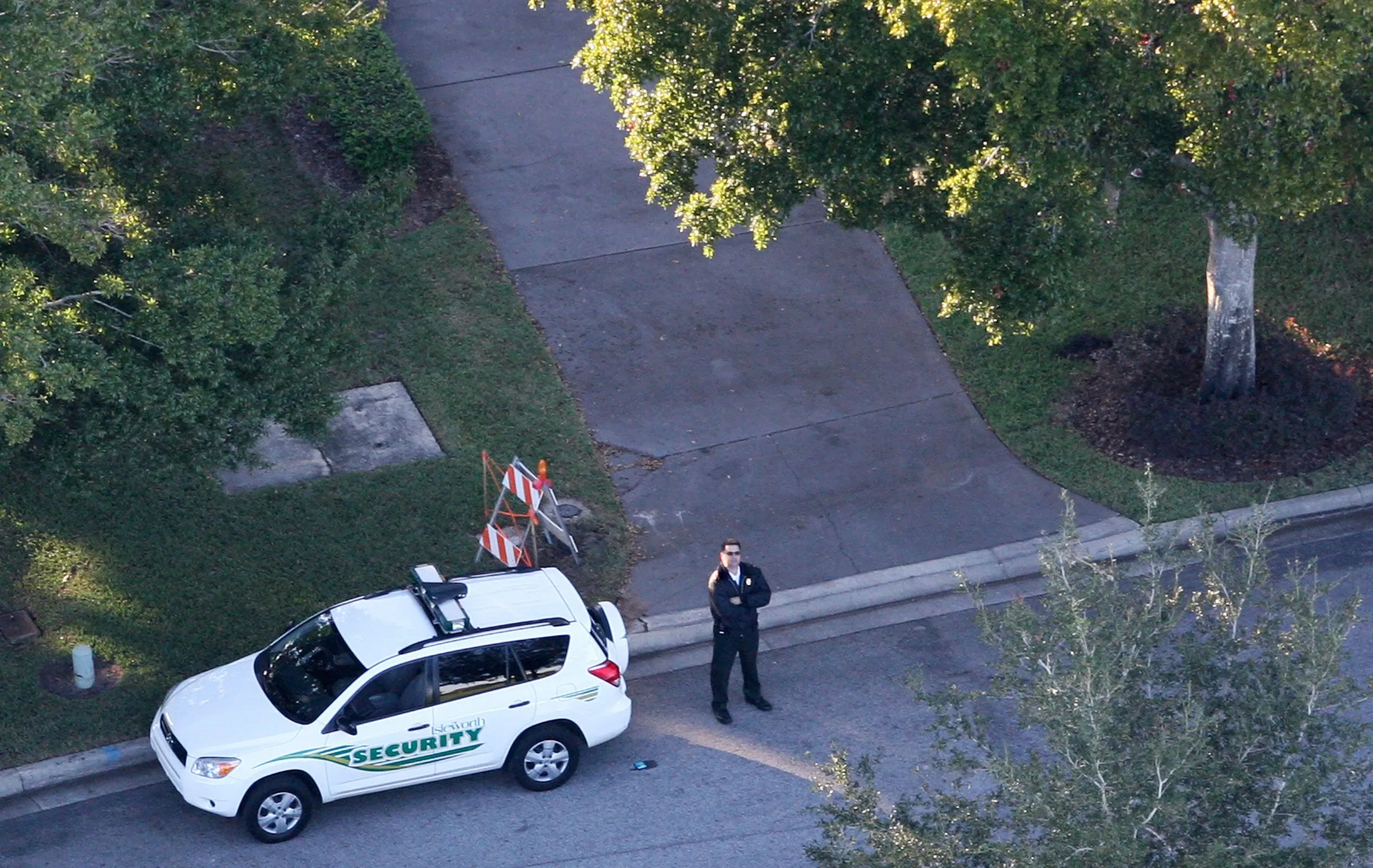 حارس أمن يقف بجانب سيارة أمنية في موقع حادث تايجر وودز، مع وجود علامات تحذيرية على الرصيف وأشجار محيطة.