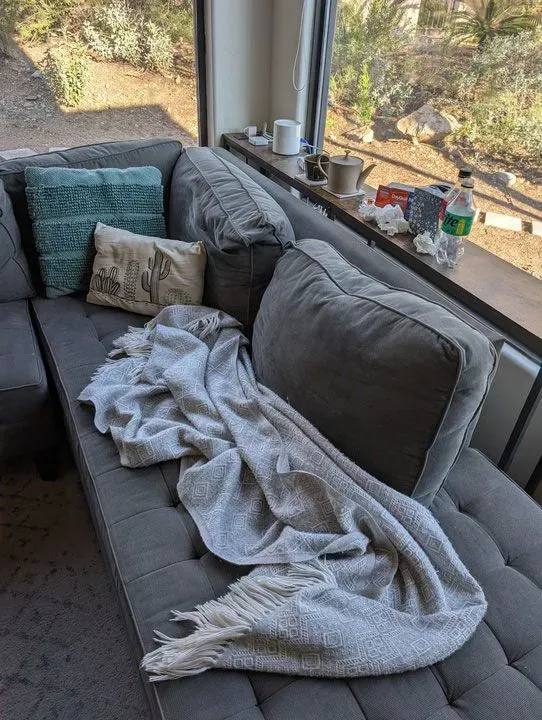 أريكة رمادية في غرفة معيشة، مغطاة ببطانية بيضاء، مع وسائد وزجاجات فارغة، تعكس أجواء المرض والإرهاق خلال موسم الإنفلونزا.