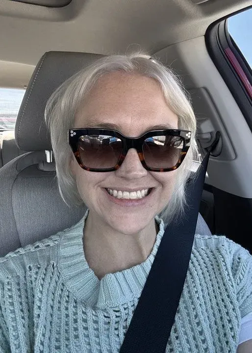 امرأة ذات شعر فاتح ترتدي نظارات شمسية وتبتسم في سيارة، تعكس شعور الأمل بعد التعافي من الإنفلونزا.