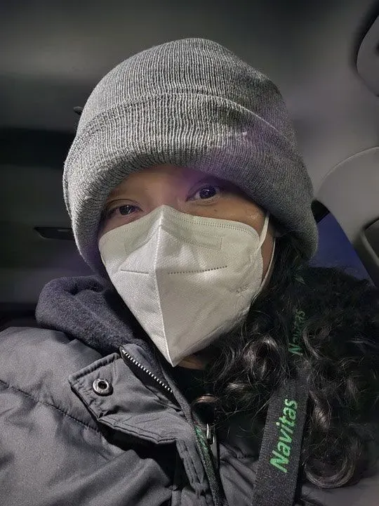 امرأة ترتدي قبعة وسماعة وجه، تجلس في السيارة، تعكس ملامحها التعب بسبب الإصابة بالإنفلونزا.